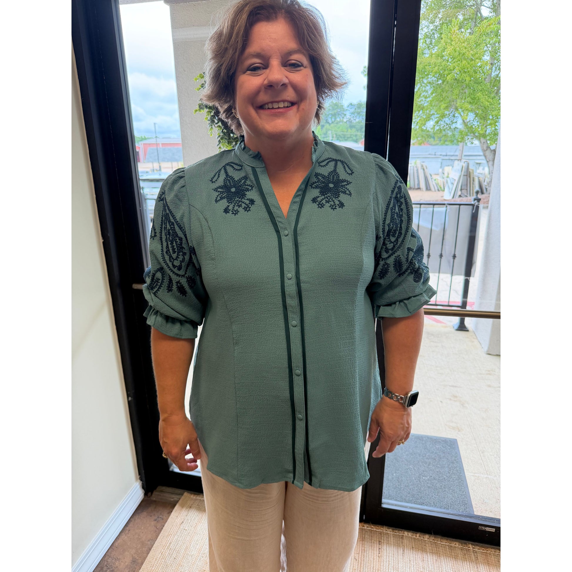 A woman wearing a sage green button-up blouse with black floral embroidery on the chest and sleeves, paired with beige pants, standing in front of a glass door.