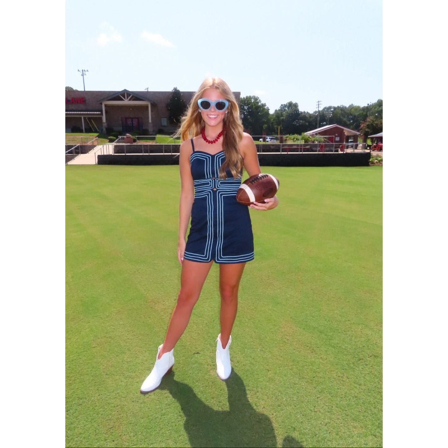 A woman stands on a grassy field holding a football, wearing a navy blue sleeveless romper with white geometric stripes, white ankle boots, and sunglasses.