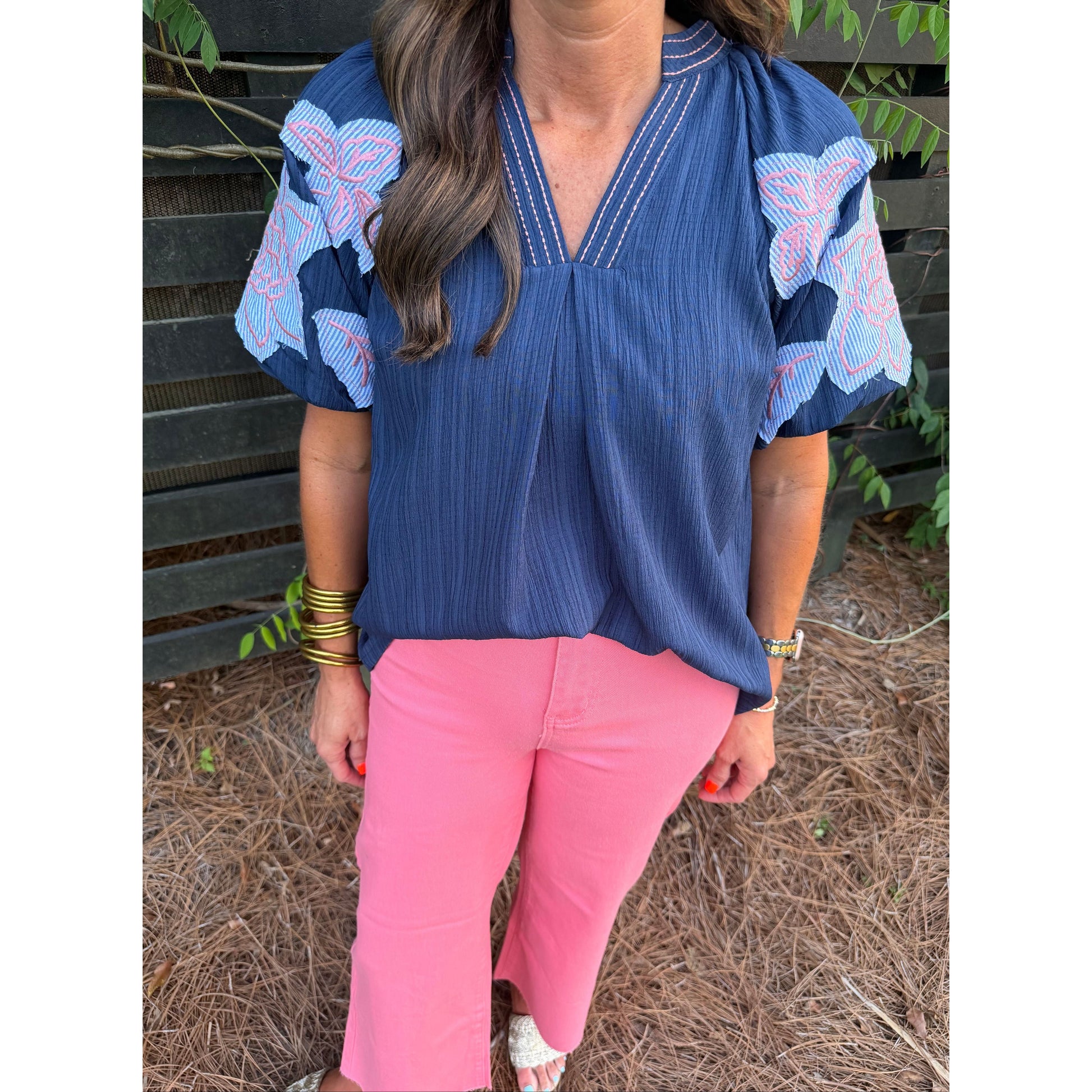 Woman wearing a blue blouse with floral patterns and pink pants standing outdoors.