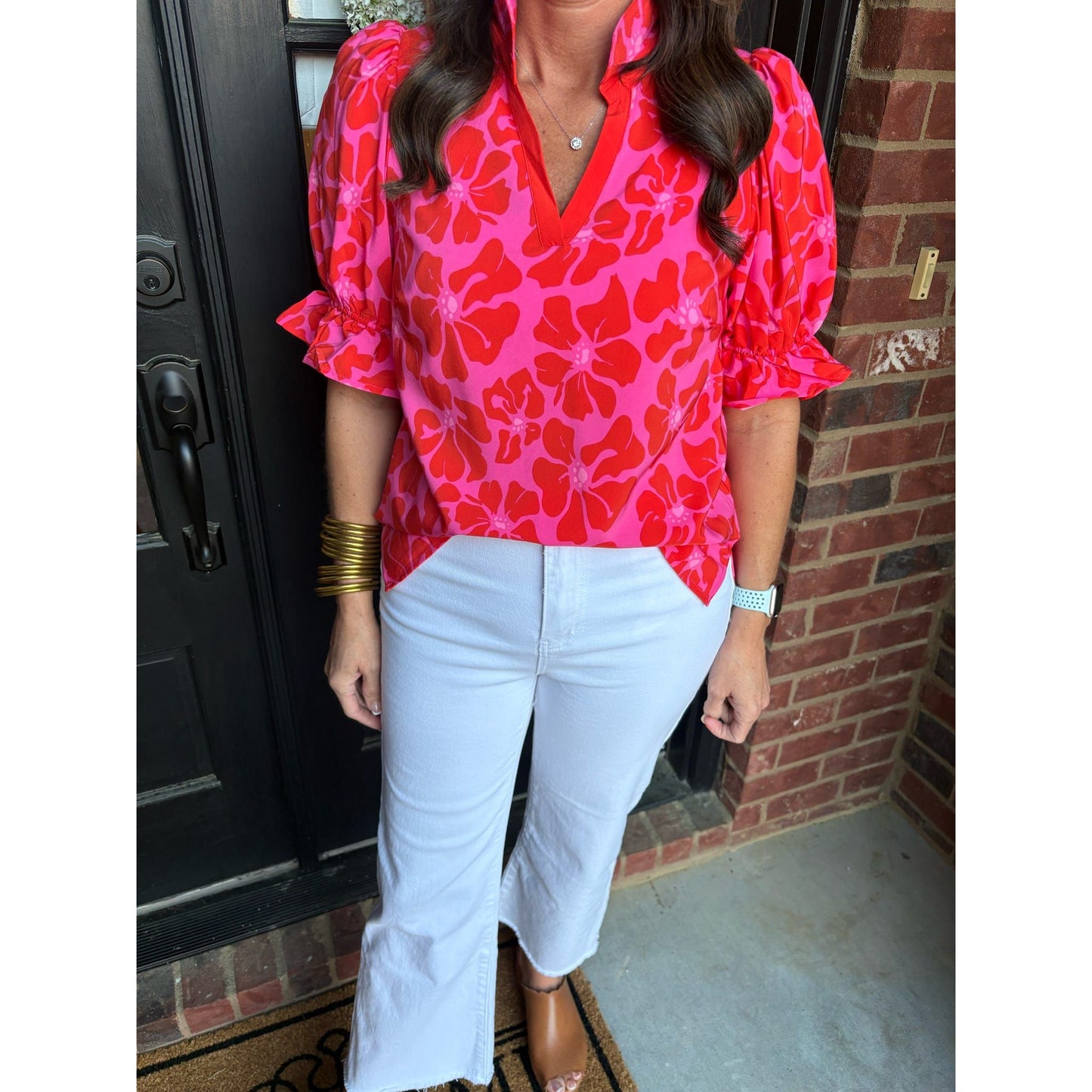 Person wearing a bright pink floral blouse and white pants standing in front of a brick wall.