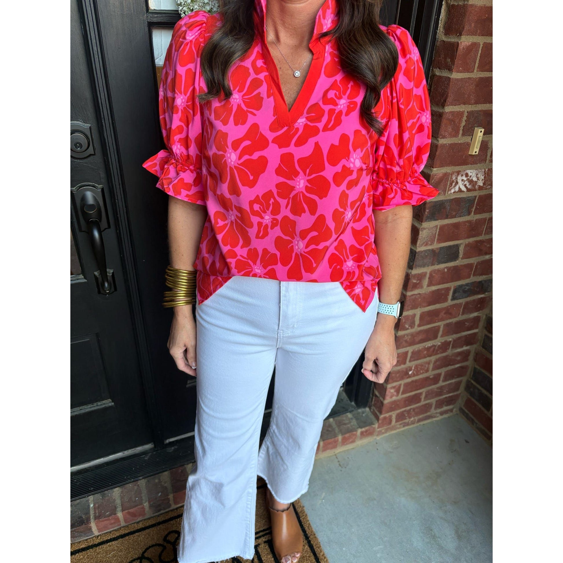 Person wearing a bright pink floral blouse and white pants standing in front of a brick wall.