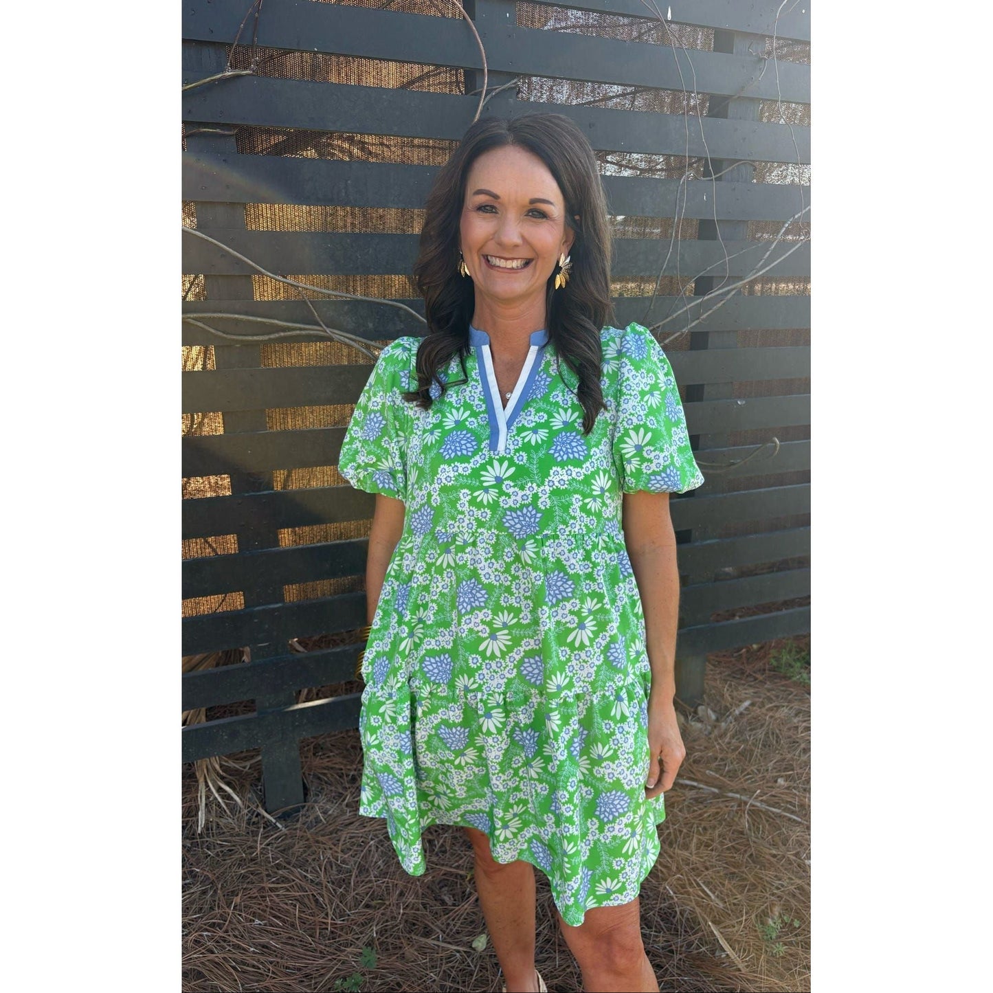 Woman wearing a green floral dress standing in front of a wooden fence.