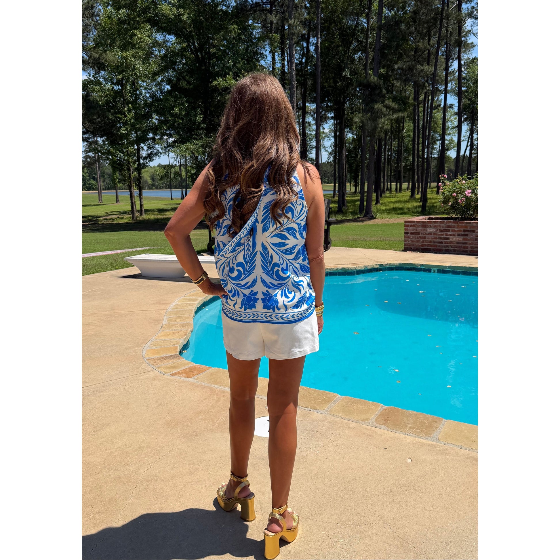 Woman in a blue and white patterned sleeveless top and white shorts standing by a pool with trees in the background.