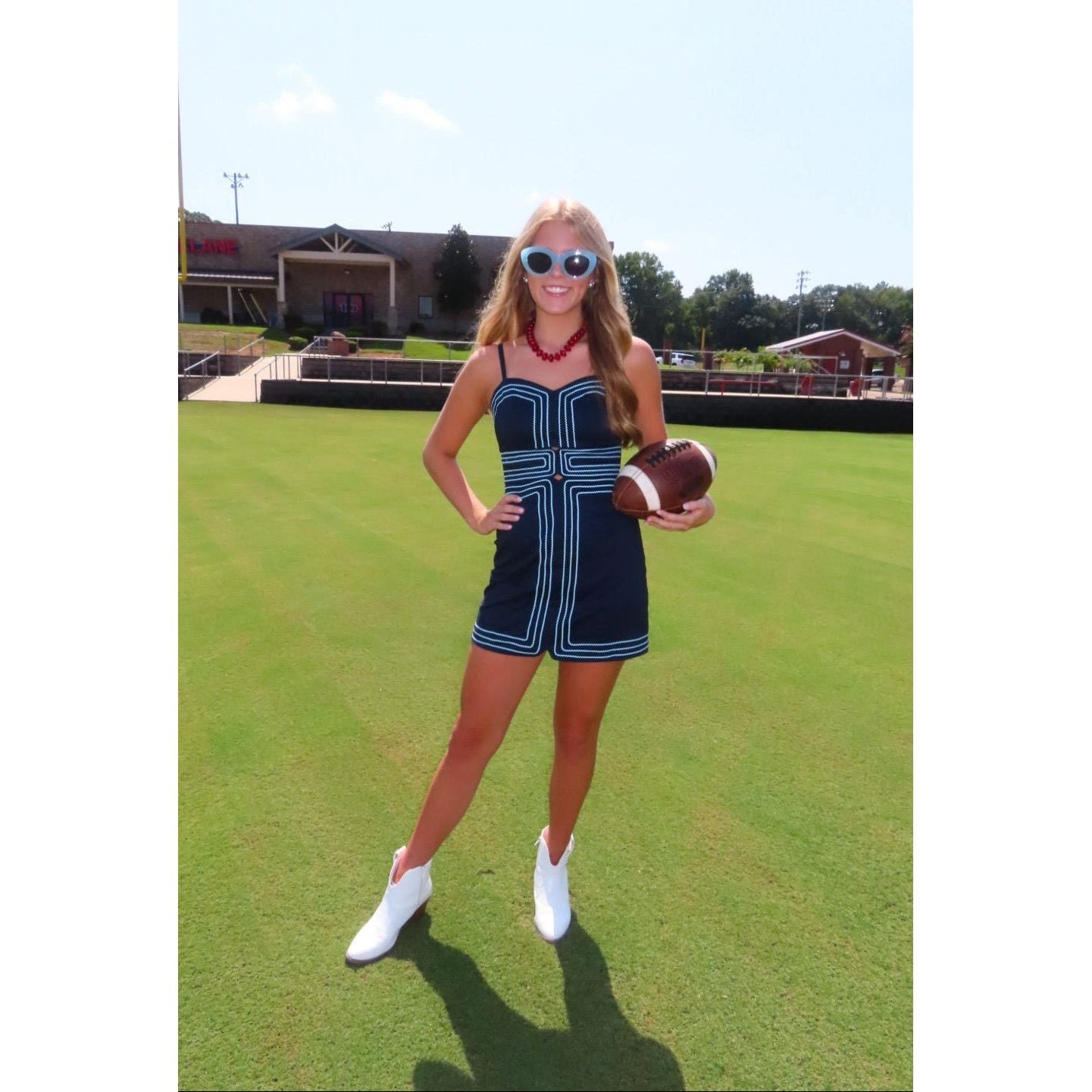 A woman stands on a grassy field holding a football, wearing a navy blue mini dress with white trim, white cowboy boots, and sunglasses.