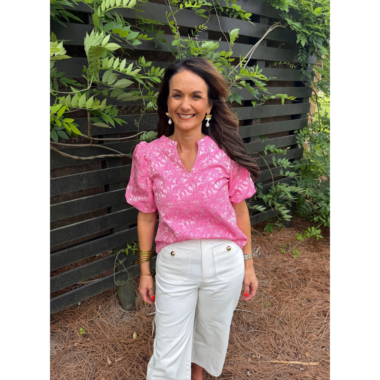 Woman in pink top and white pants standing outdoors with plants and a bench in the background