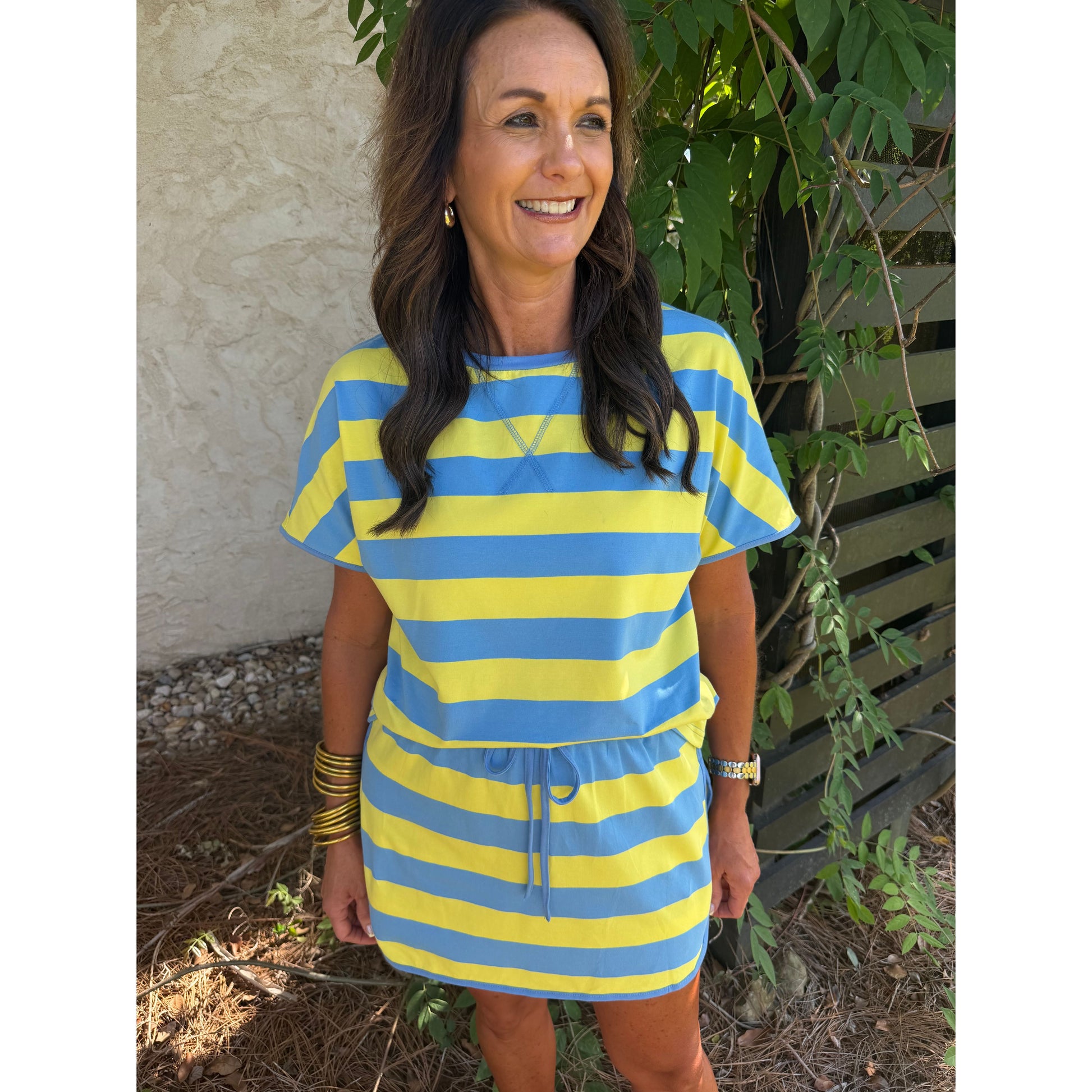 Woman wearing a blue and yellow striped dress standing outdoors with greenery around.