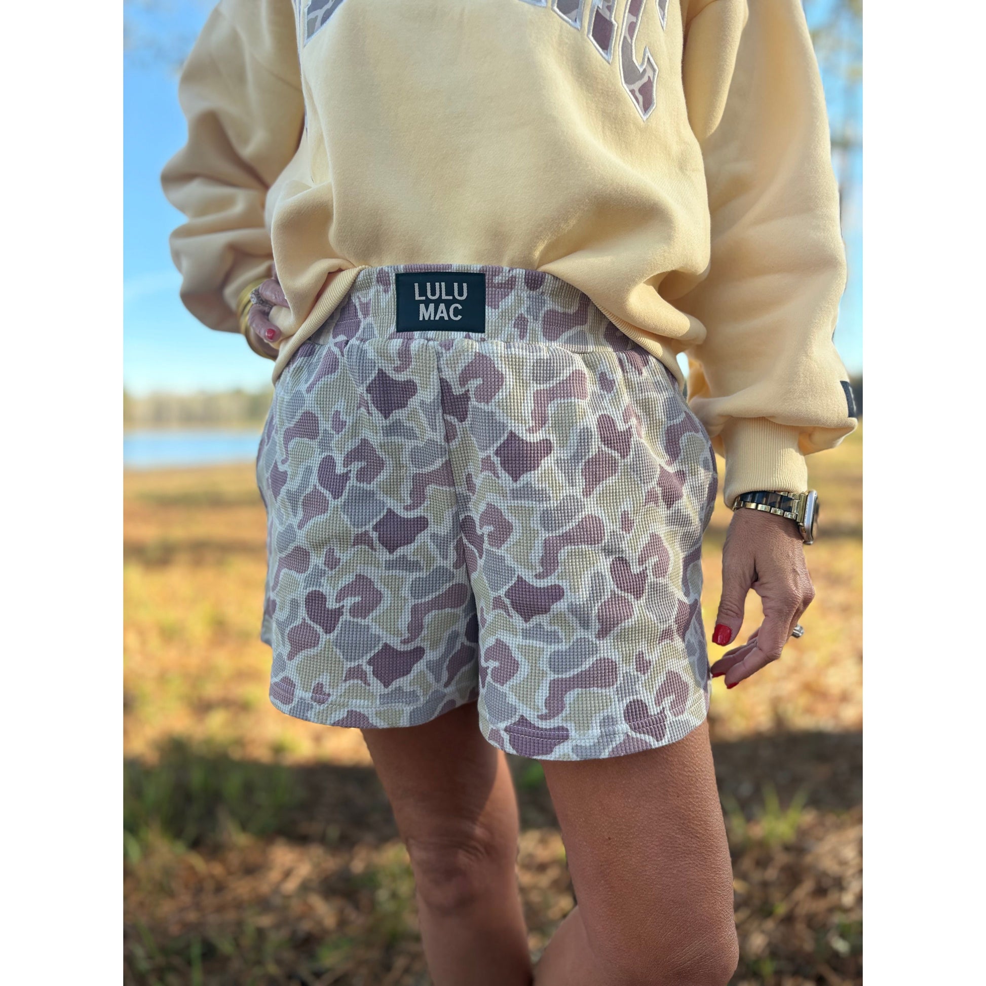 Person wearing a beige sweatshirt and patterned shorts with LULU MAC branding outdoors.