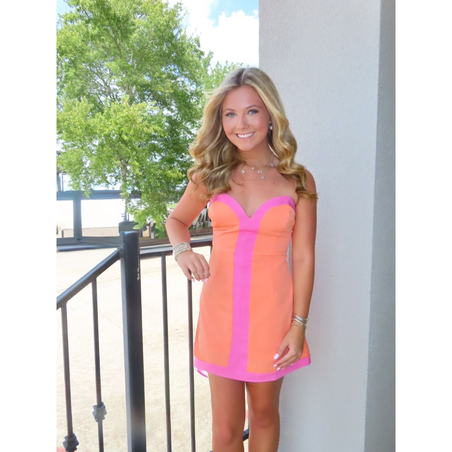 A woman modeling a short, sleeveless corset-style dress with a bright coral-orange body and vivid pink trim, standing on an outdoor balcony with trees and a white wall in the background.