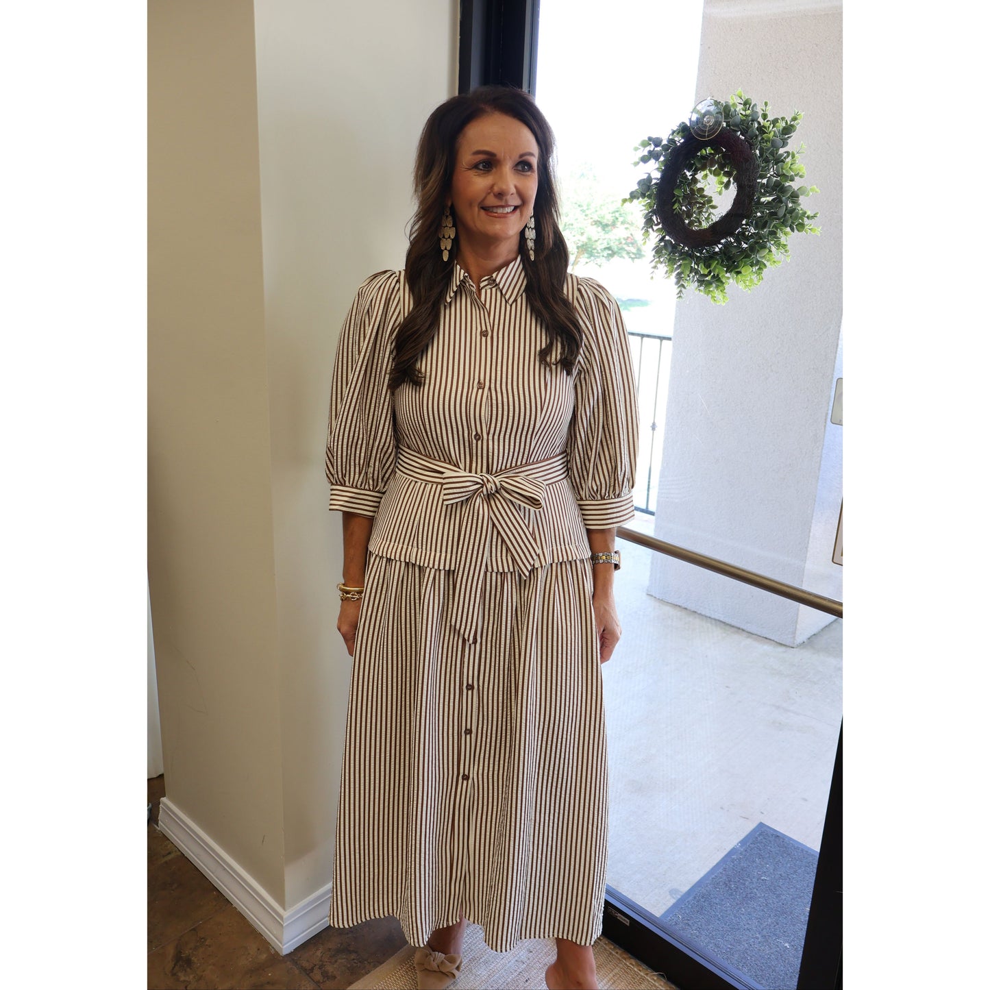 A woman wearing a beige and brown vertically striped midi shirt dress with puff sleeves, a button-down front, and a matching fabric tie belt at the waist, standing in an indoor setting.