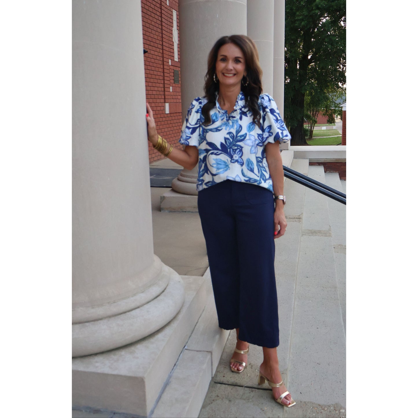 Woman in a blue floral blouse and navy pants standing outdoors.
