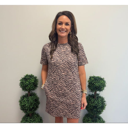 A woman wearing a short-sleeve brown and beige zebra-print dress, standing indoors between two green topiary trees against a plain wall.