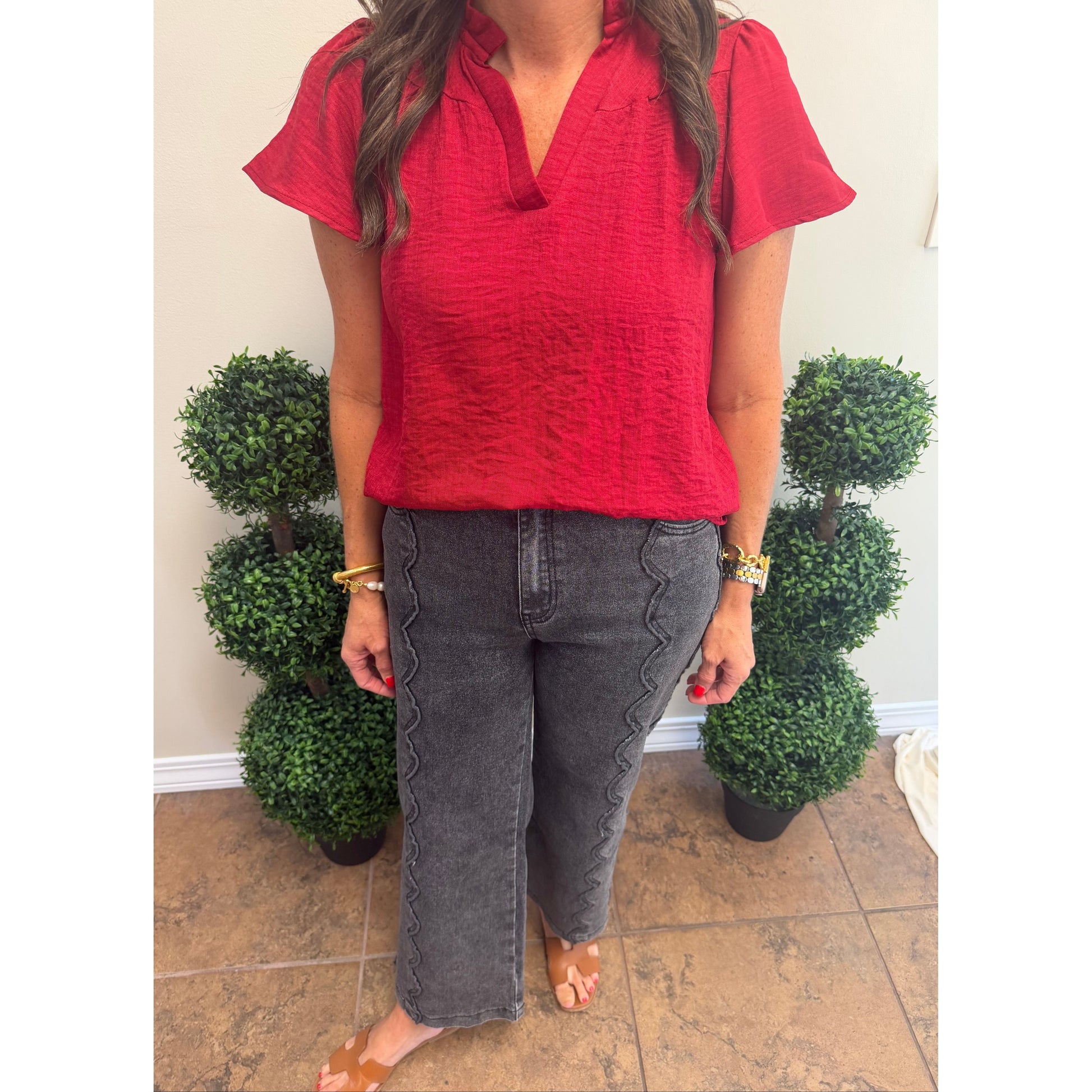 A woman wearing a textured red short-sleeve collared top and dark gray cropped jeans with decorative zigzag stitching down the sides, standing indoors between two potted topiary trees.