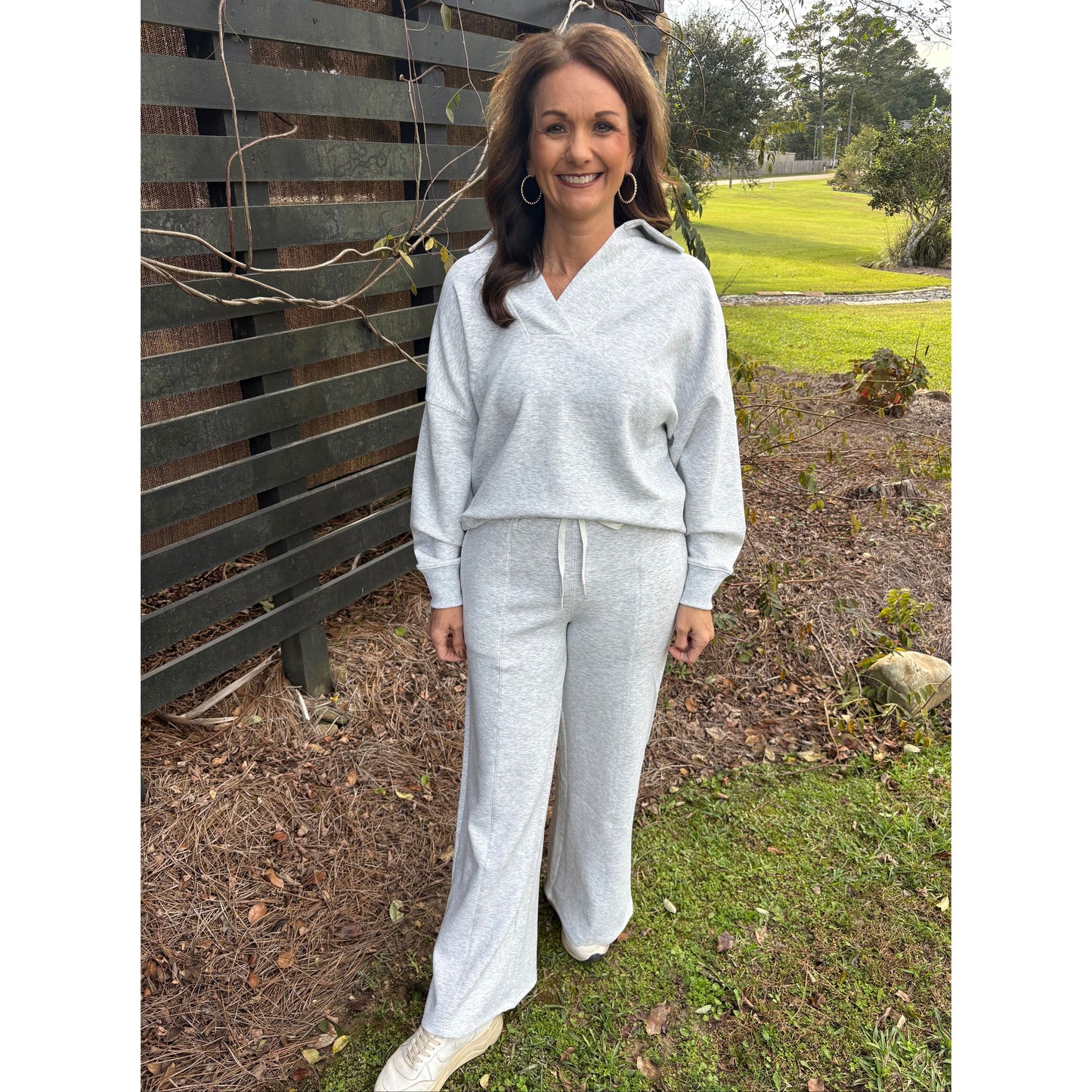 Woman in a light gray outfit standing outdoors on a wooden bench with grass and trees in the background.
