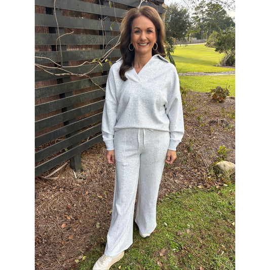 Woman in a light gray outfit standing outdoors on a wooden bench with grass and trees in the background.