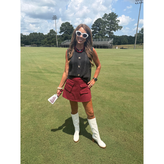 A woman stands on a grassy field wearing a sleeveless black knit top with gold buttons, a red mini skirt with large front pockets, white cowboy boots, and white sunglasses. She is holding a white bottle with red text.