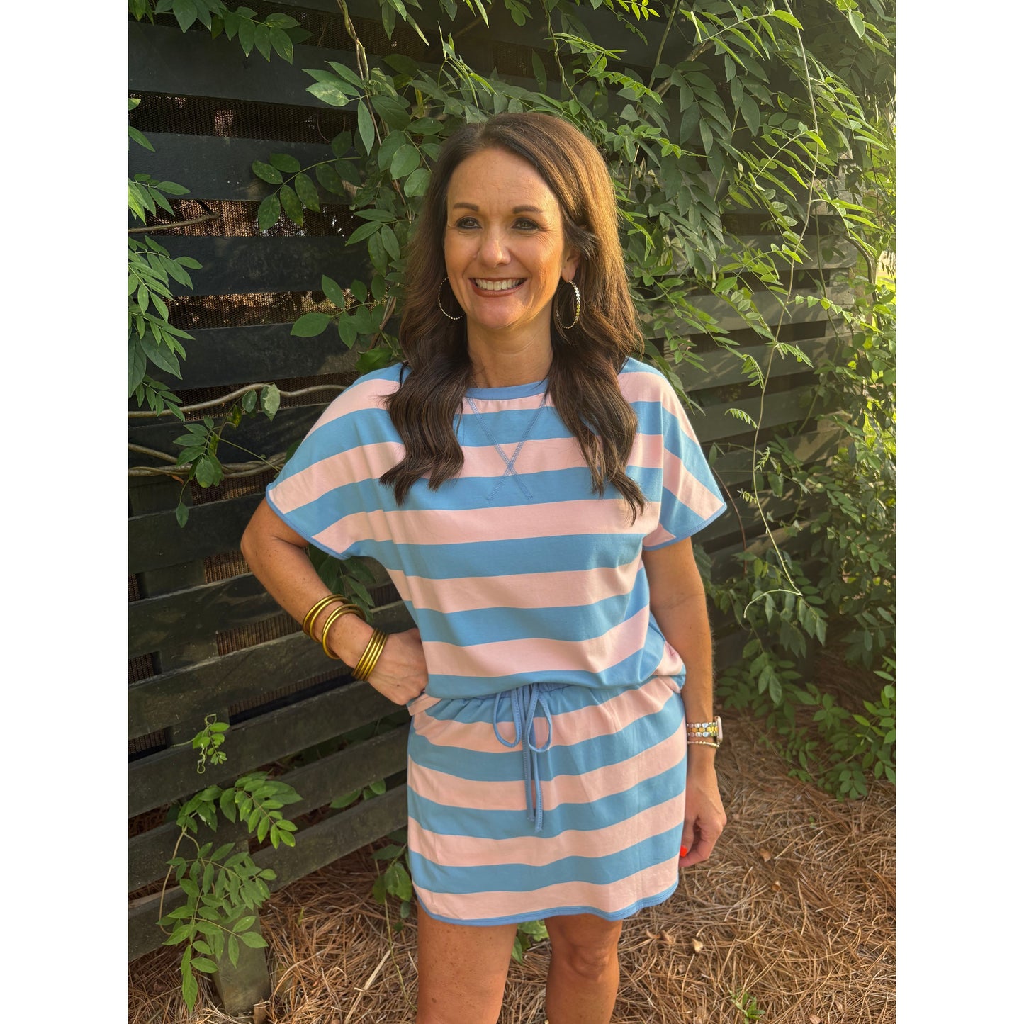 Woman wearing a pink and blue striped dress standing outdoors with greenery around.