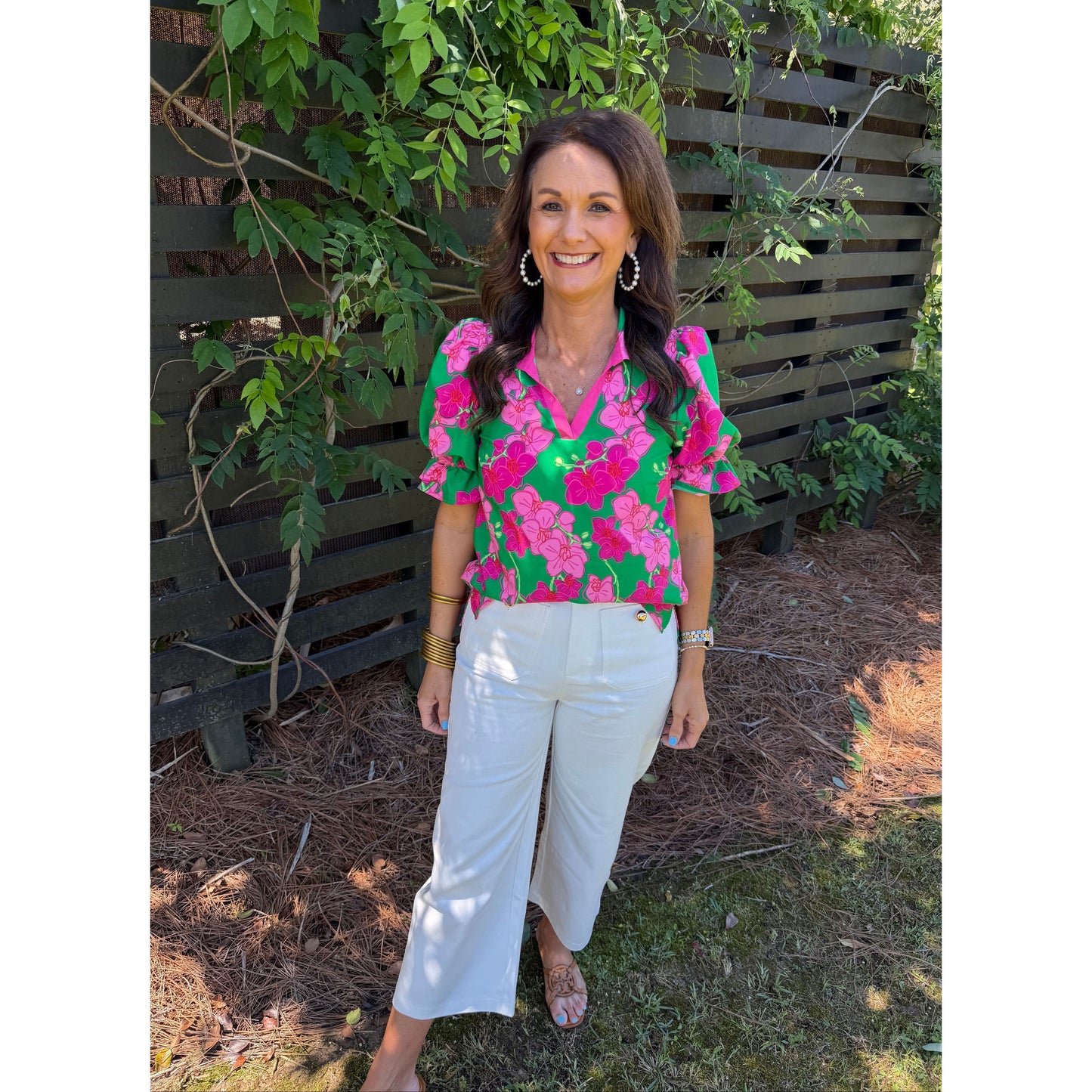 Person wearing a colorful floral blouse and white pants standing outdoors with greenery around.