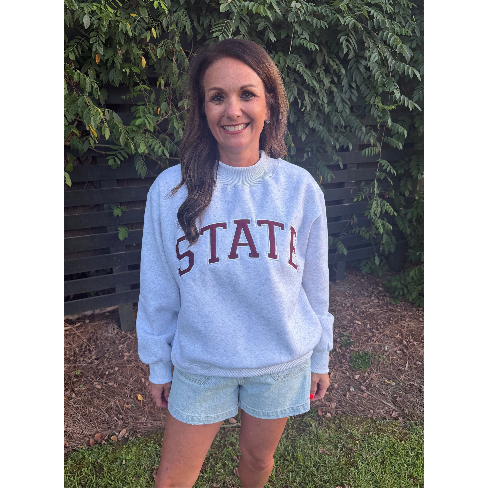A woman wearing a light blue sweatshirt with 'STATE' printed in dark red across the chest, paired with denim shorts, standing outdoors on a bench with greenery and mulch in the background.