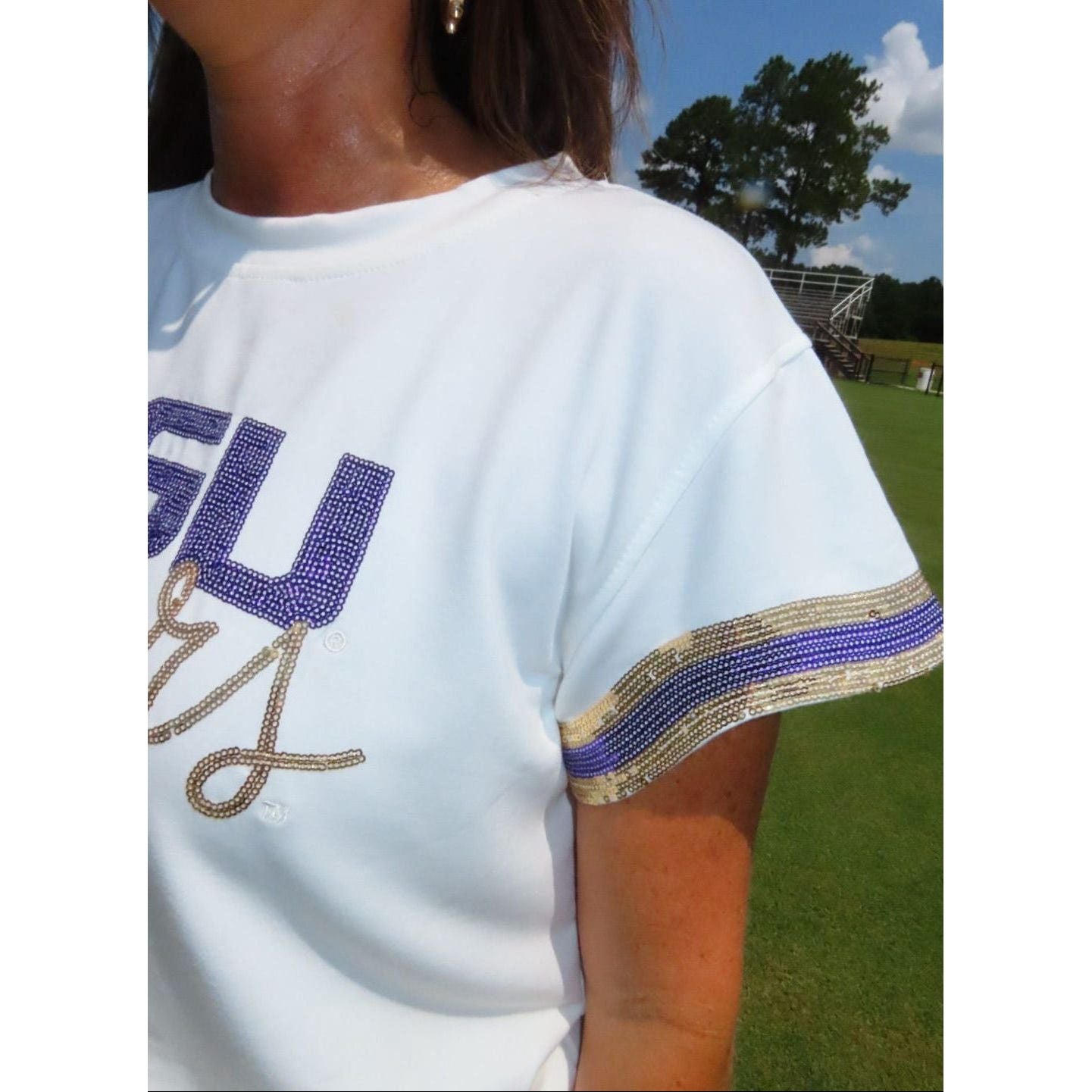 A close-up of a white short-sleeve t-shirt with a purple and gold rhinestone 'LSU Tigers' graphic on the chest and matching striped trim on the sleeve, worn by a model outdoors on a grassy field.