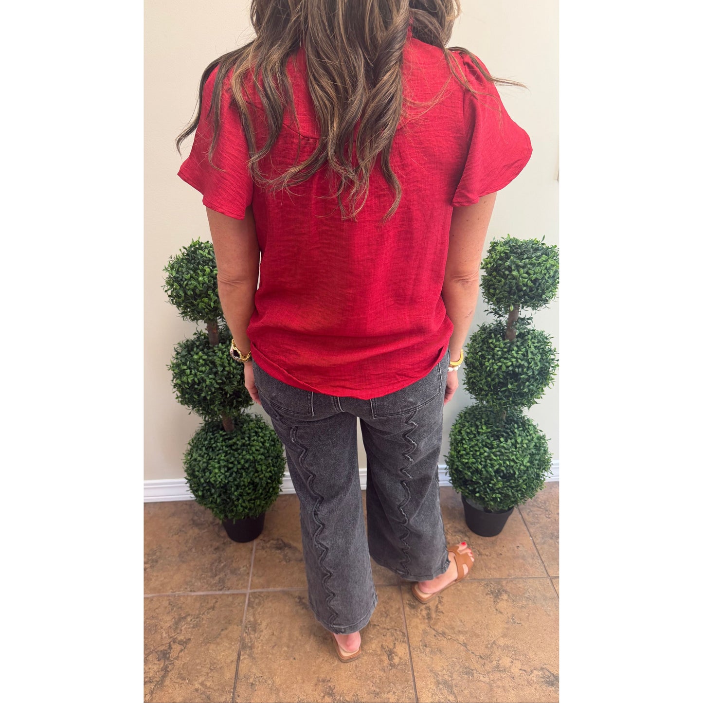 A woman viewed from behind wearing a bright red short-sleeve top and dark gray wide-leg jeans, standing indoors between two green potted topiary trees against a beige wall.