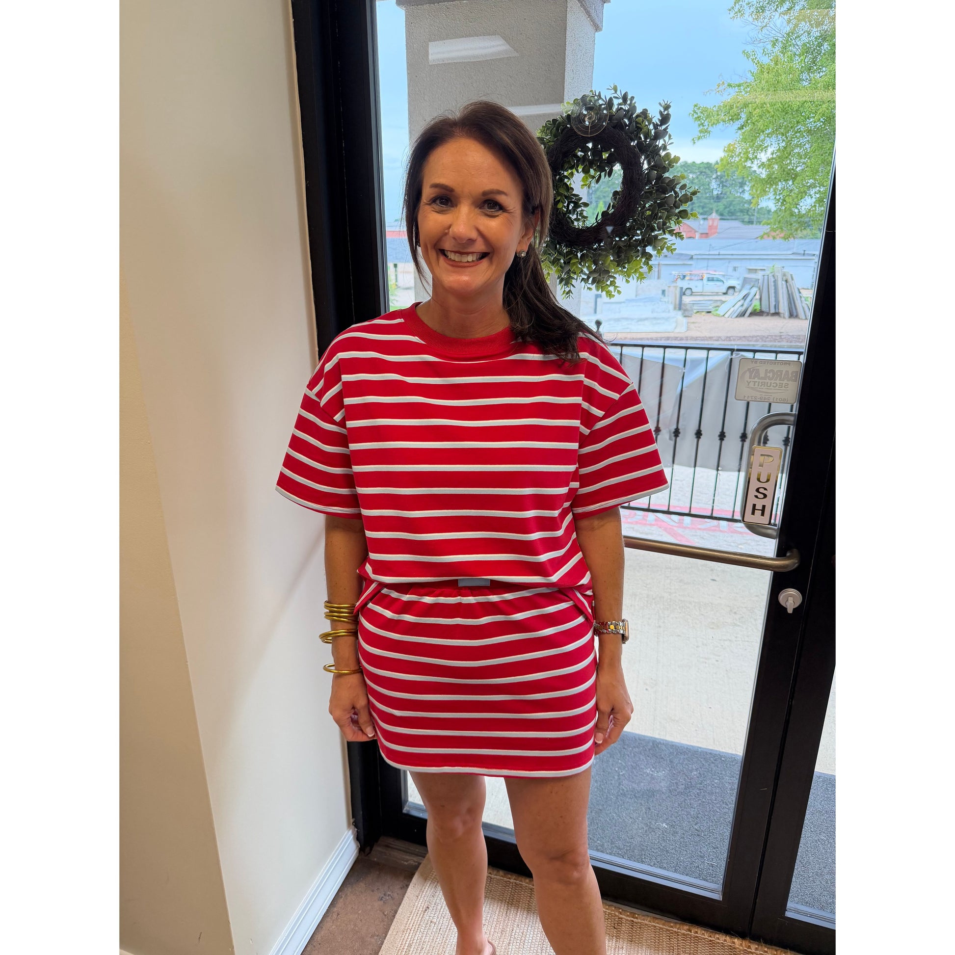 A woman wearing a red and white horizontally striped short-sleeve t-shirt and matching striped mini skirt, standing in front of a glass door with a storefront and outdoor scene visible behind her.
