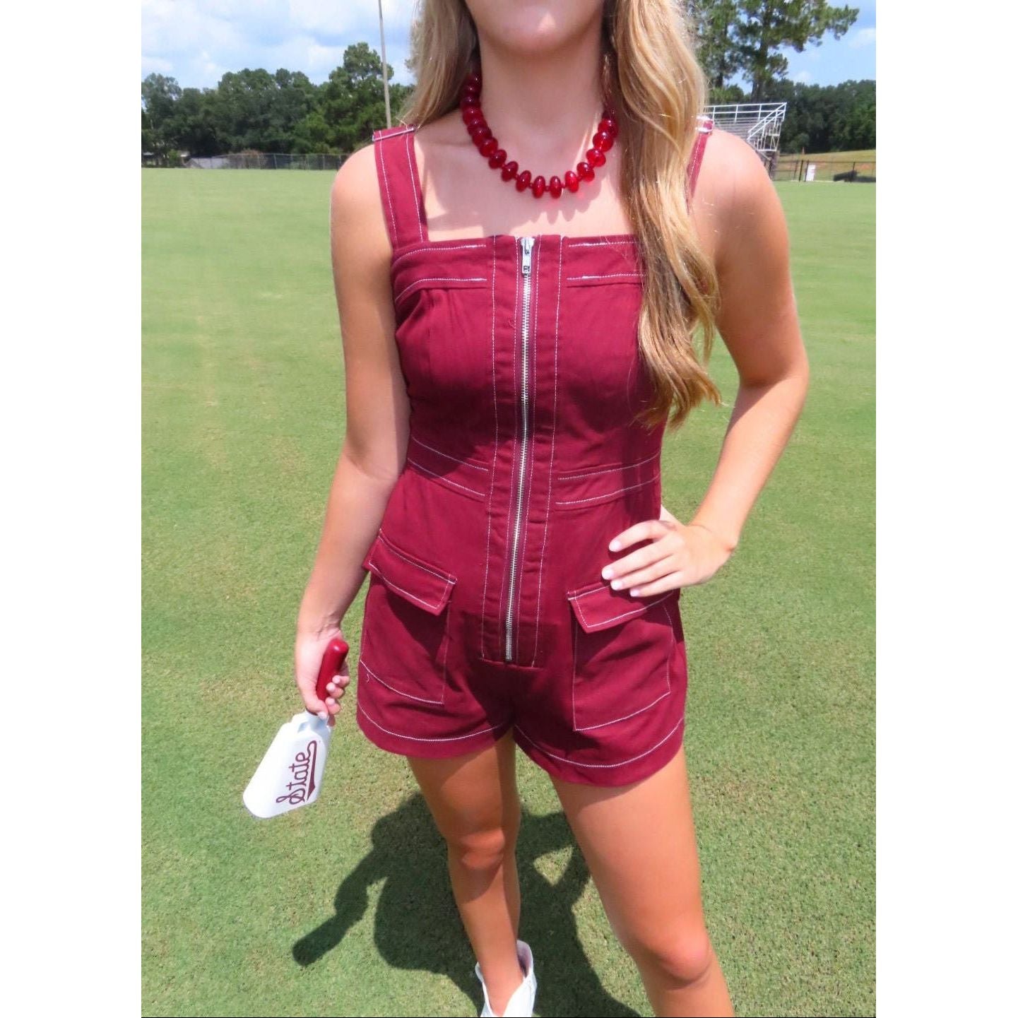 A woman wearing a sleeveless burgundy romper with a front zipper and contrast stitching, standing on a green grass field.