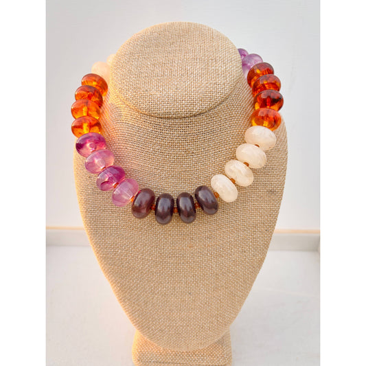 A beaded necklace displayed on a beige fabric-covered jewelry bust. The necklace features translucent amber-orange beads, pink-purple beads, dark brown beads, and cream-colored beads, arranged in a gradient pattern.