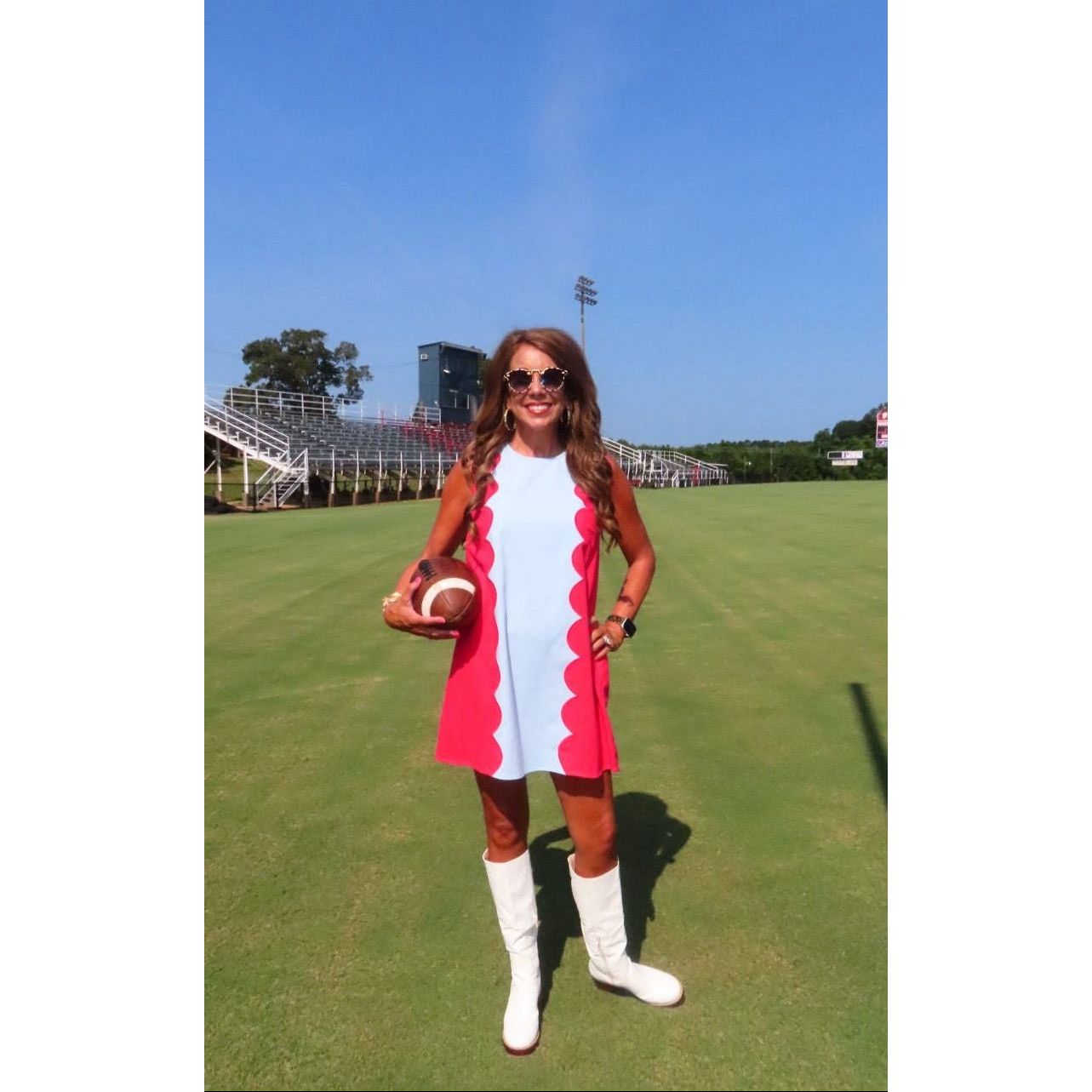 A woman stands on a grassy field holding a football, wearing a retro-style red and white dress with scalloped edges, white knee-high boots, and sunglasses. Stadium bleachers and a blue sky are visible in the background.