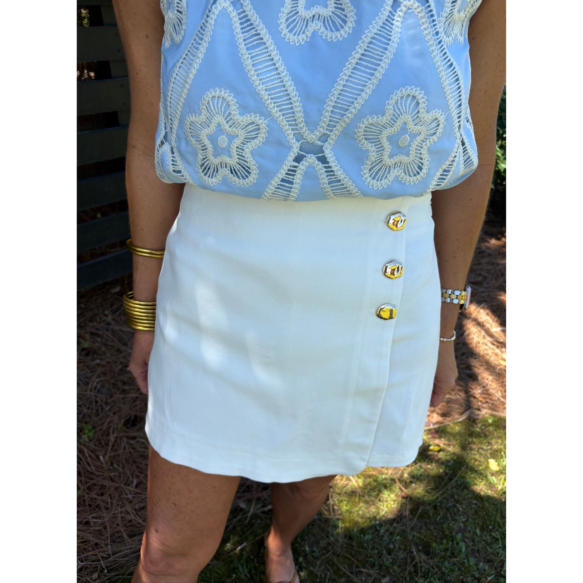 Person wearing a light blue embroidered top and white skirt with yellow buttons outdoors.