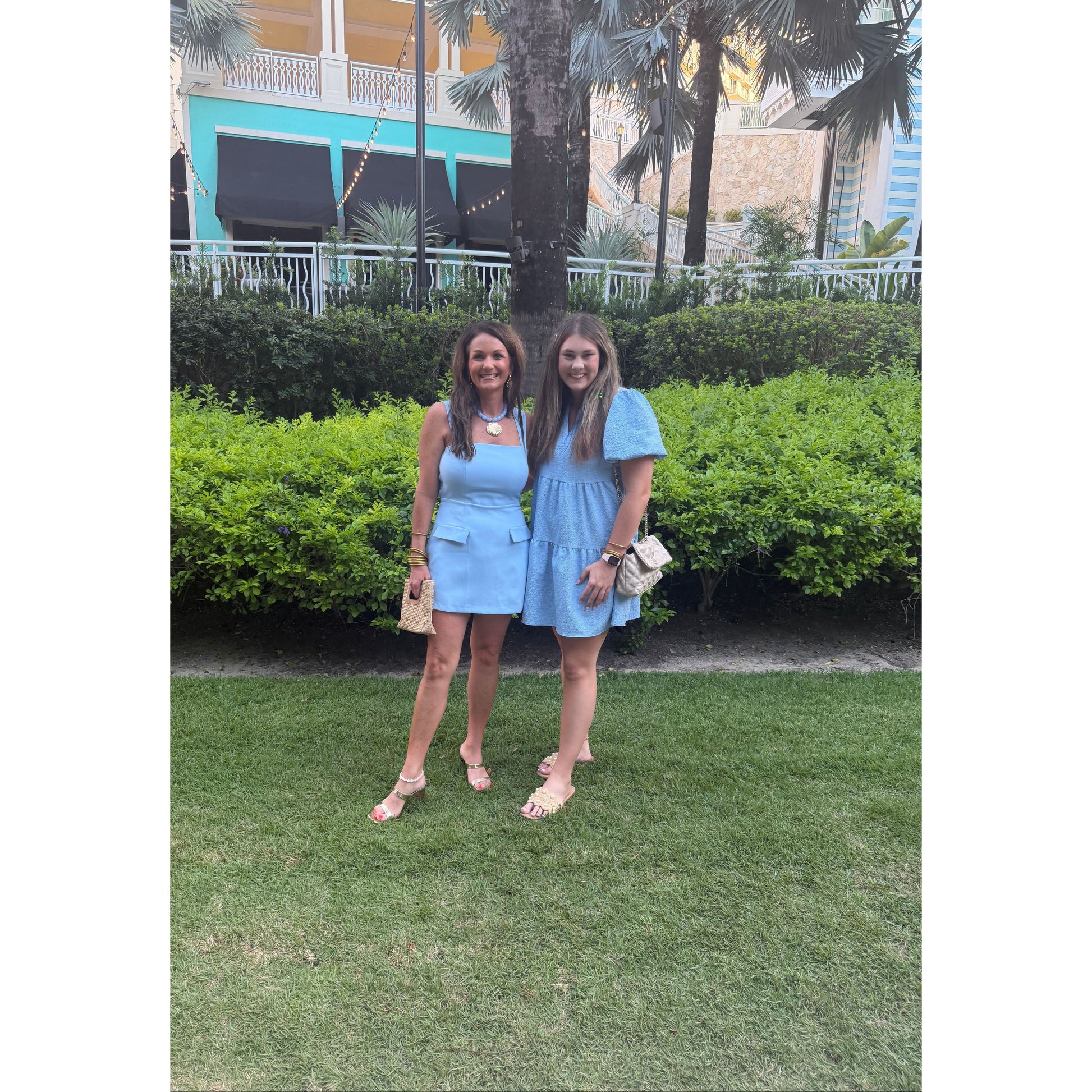 Two women standing on a green lawn wearing matching light blue mini dresses, with hedges, palm trees, and a building in the background.