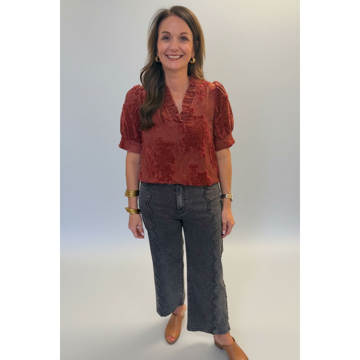 A woman wearing a textured rust-red short-sleeve blouse and dark gray cropped jeans with scalloped edges, standing in a home interior next to a white column.