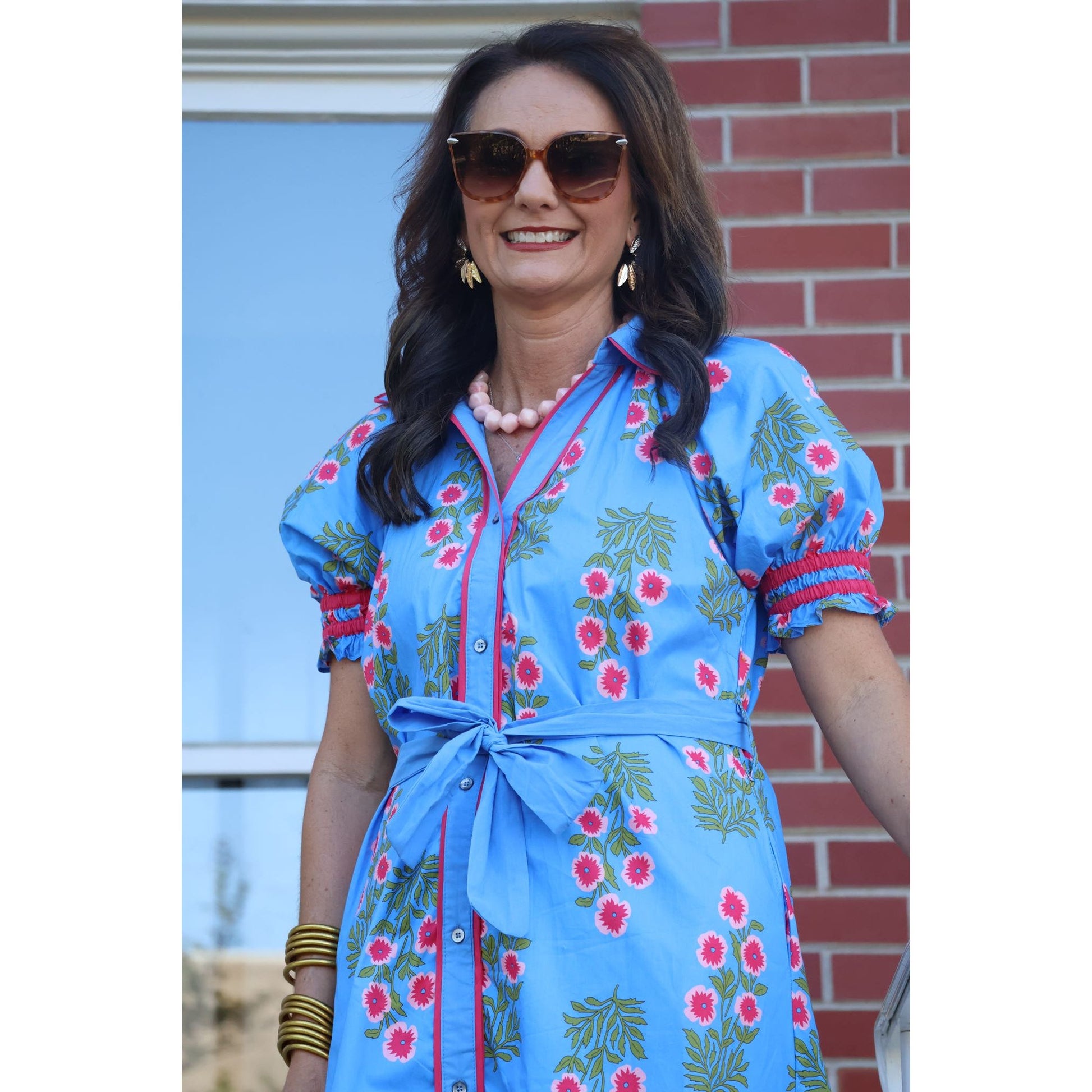 Woman wearing a blue floral dress with sunglasses and earrings, standing in front of a brick wall.