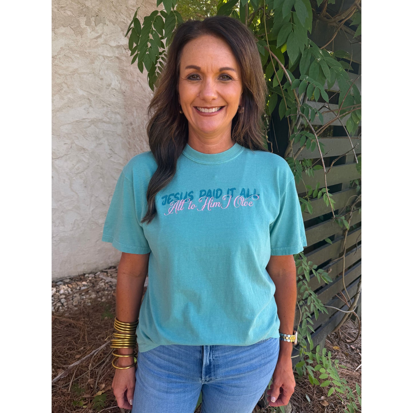 Woman wearing a blue t-shirt with text, standing outdoors against a wall and plants.