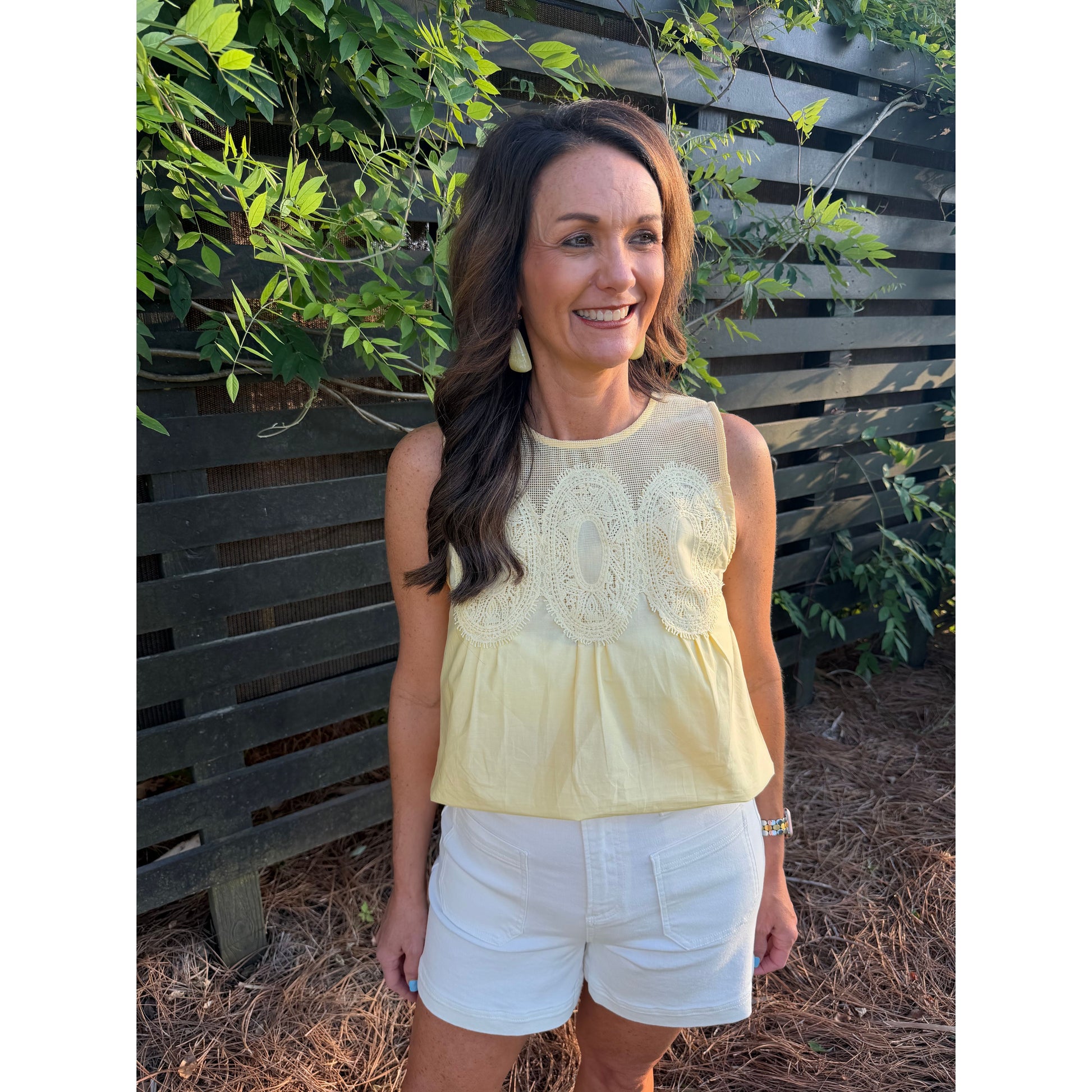 Woman wearing a yellow top and white shorts standing outdoors with greenery in the background