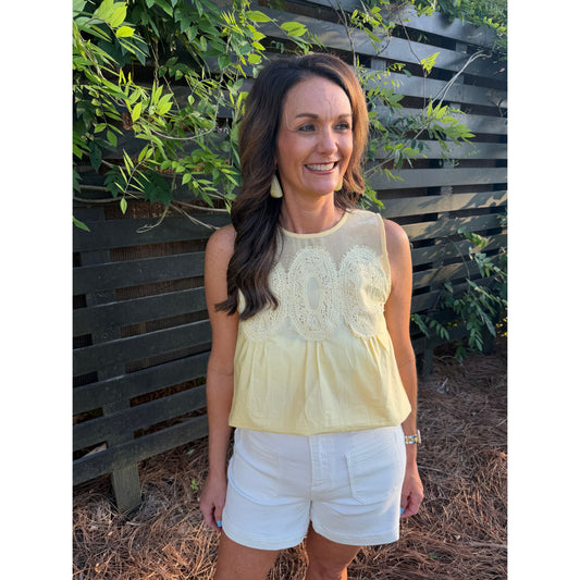Woman wearing a yellow top and white shorts standing outdoors with greenery in the background