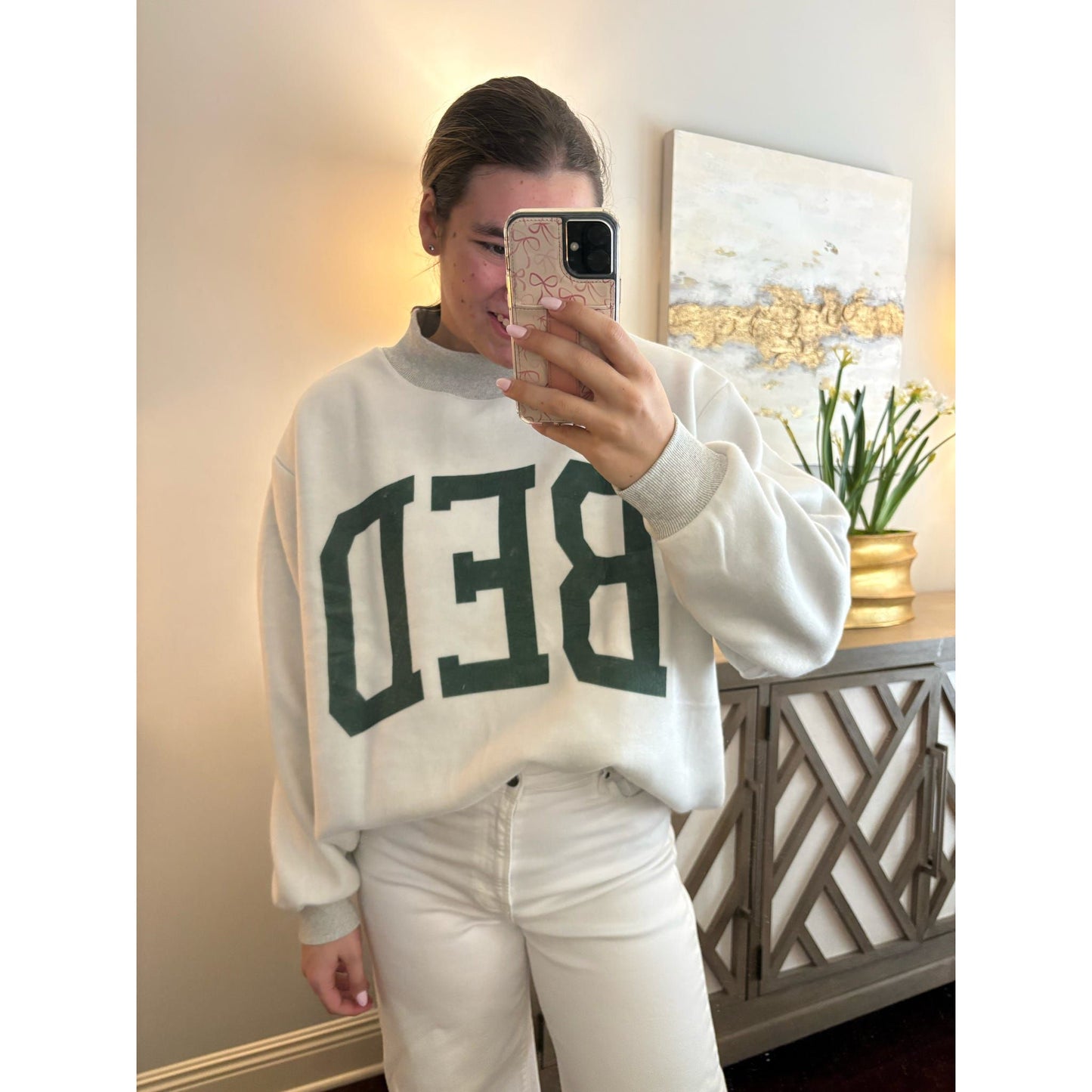 Person wearing a white sweatshirt with 'BEDE' printed on it, taking a mirror selfie.