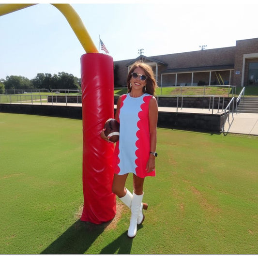 A woman wearing a light blue sleeveless dress with bright pink scalloped side panels and white knee-high boots stands on a grassy field next to a red football pylon, holding a football.