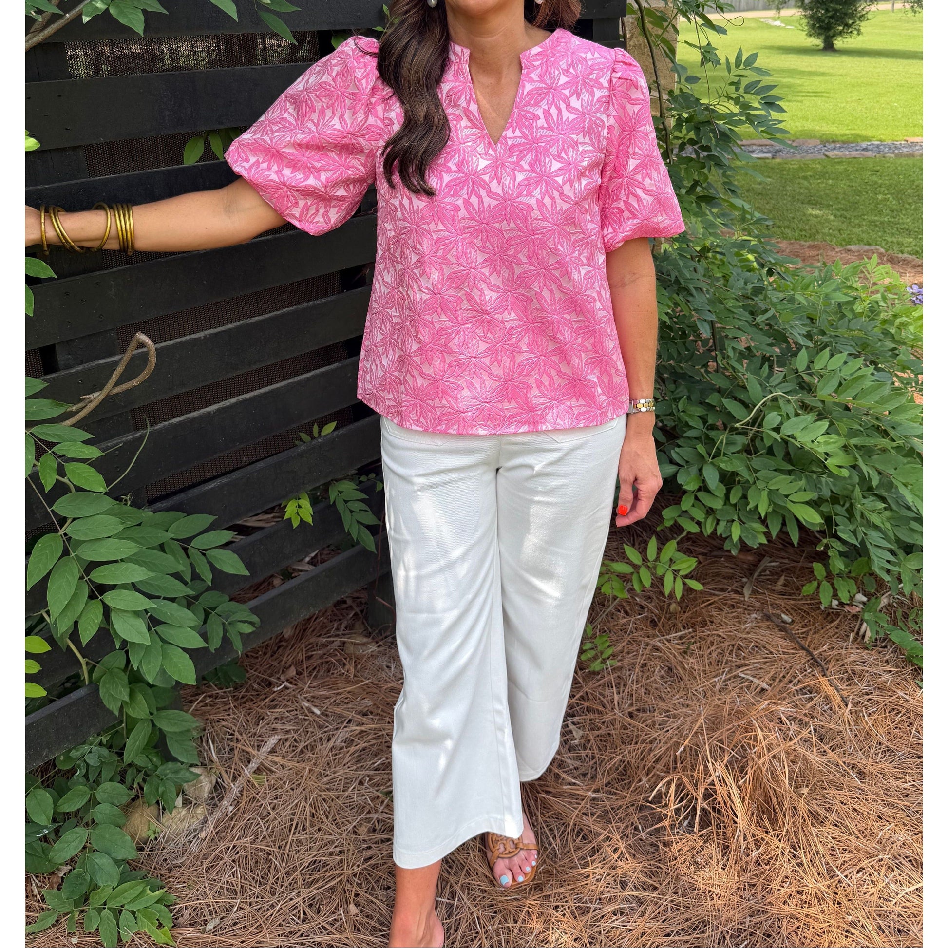 Woman in a pink top and white pants standing outdoors with greenery around