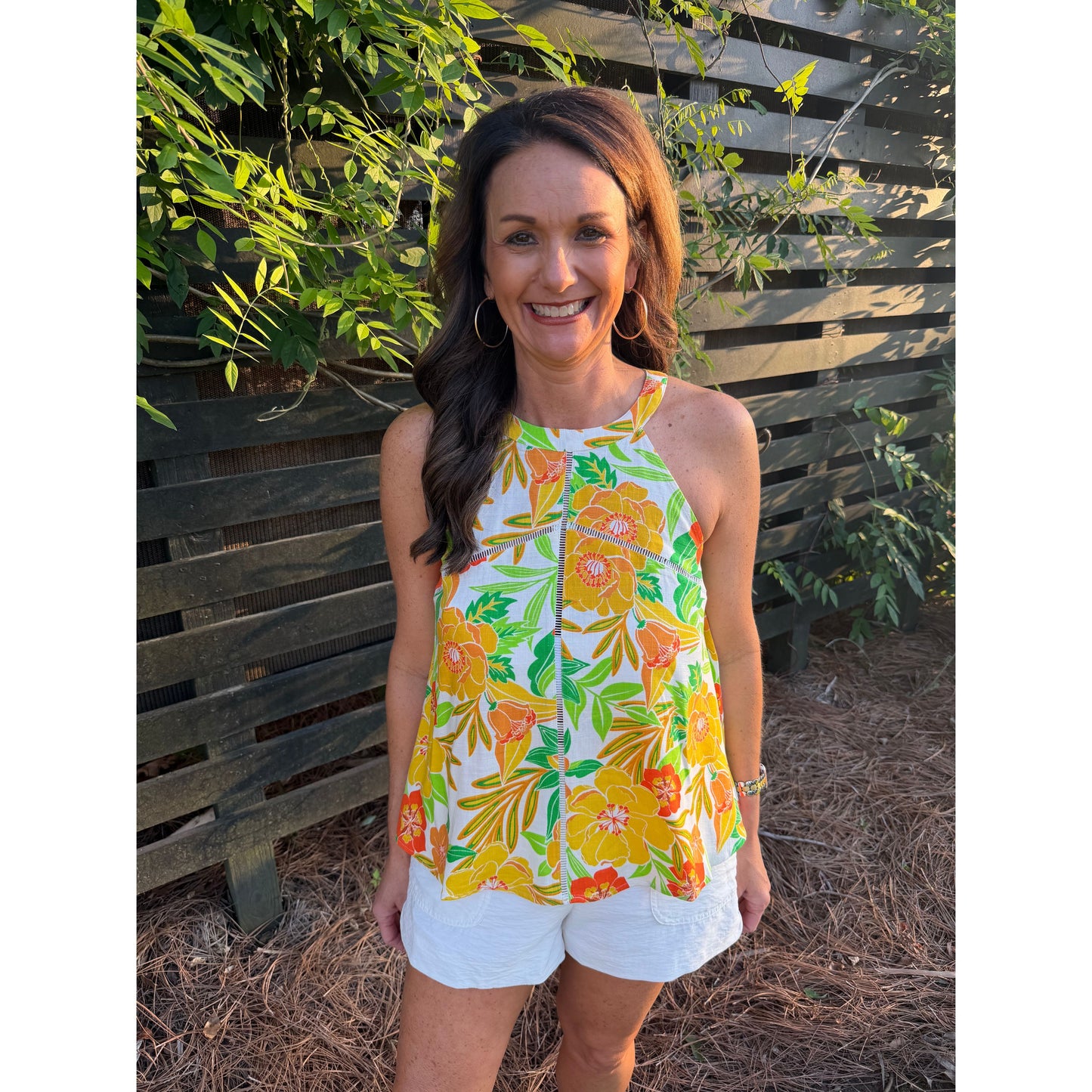 Woman wearing a colorful floral top and white shorts standing in front of a wooden fence with greenery.