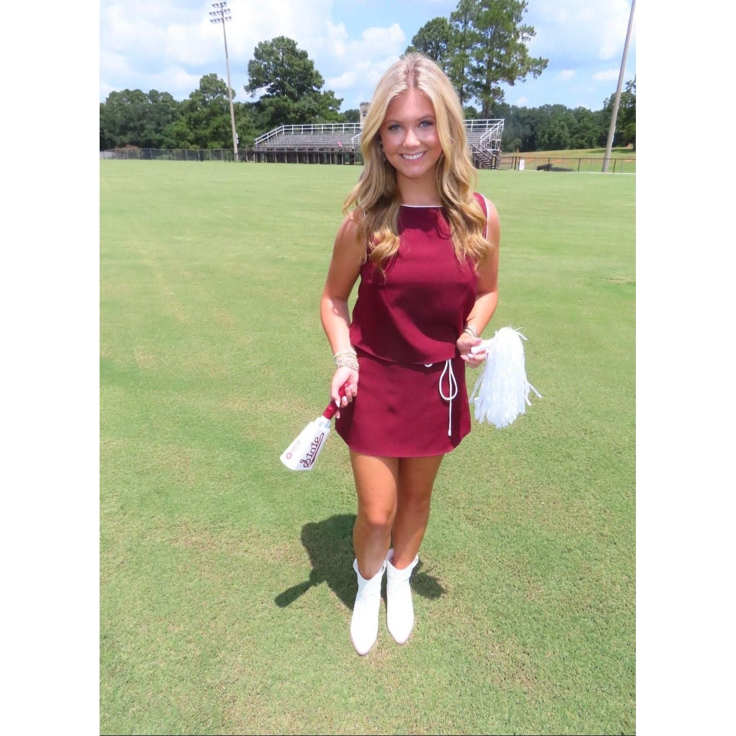 A woman stands on a grassy field wearing a sleeveless burgundy mini dress with a white drawstring, white ankle boots, and holding white pom-poms.