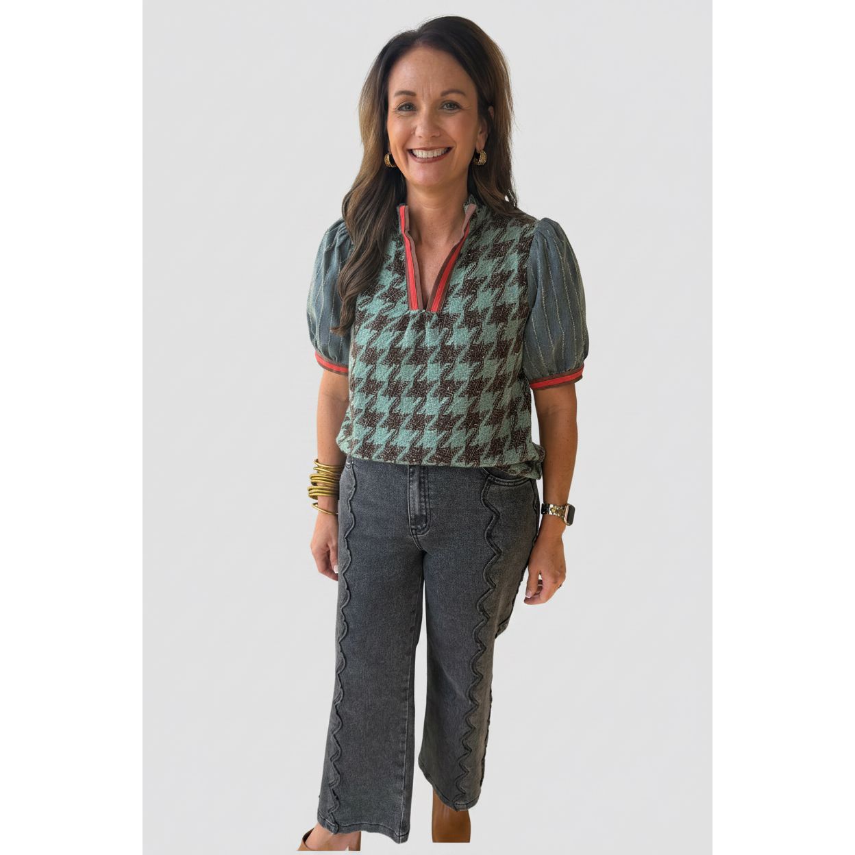 A woman stands on a covered outdoor walkway wearing a short-sleeve knit top with a green and brown houndstooth pattern, sheer blue-gray puff sleeves with red trim, and a red split neckline. She is also wearing dark gray cropped jeans with decorative ruffle detailing along the outer seams.