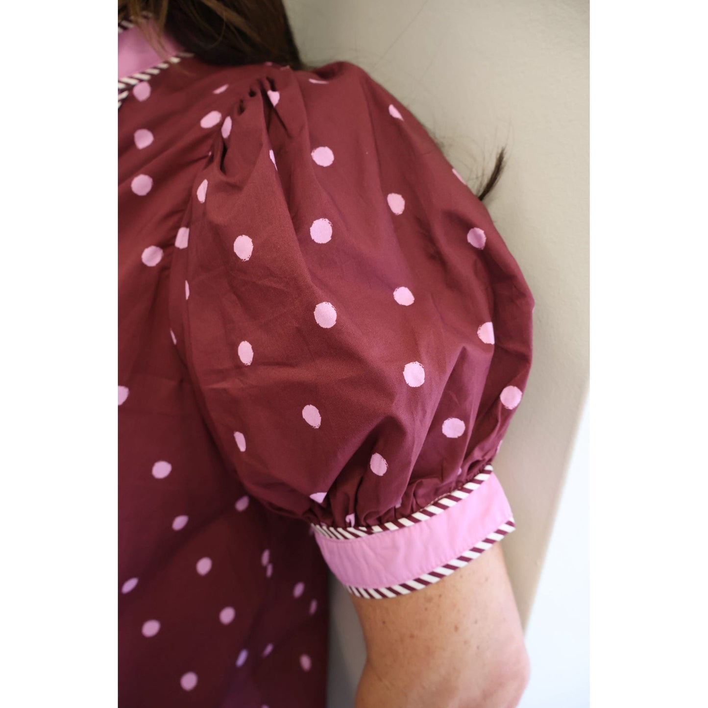Close-up of a person wearing a burgundy dress with light pink polka dots and a contrasting light pink cuff with striped trim, leaning against a plain wall.
