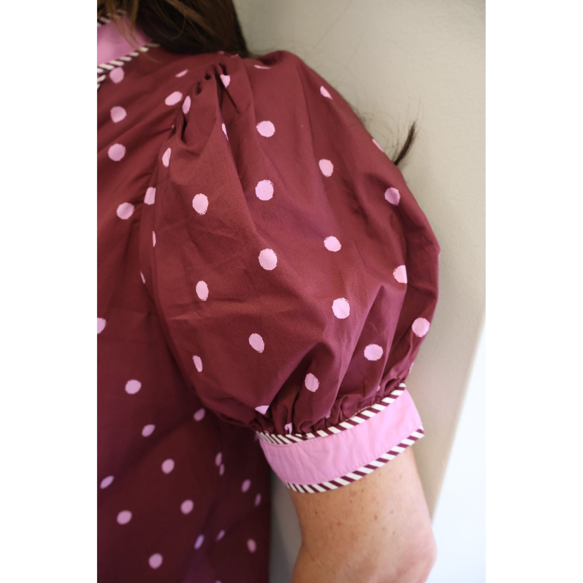 Close-up of a person wearing a burgundy dress with light pink polka dots and a contrasting light pink cuff with striped trim, leaning against a plain wall.