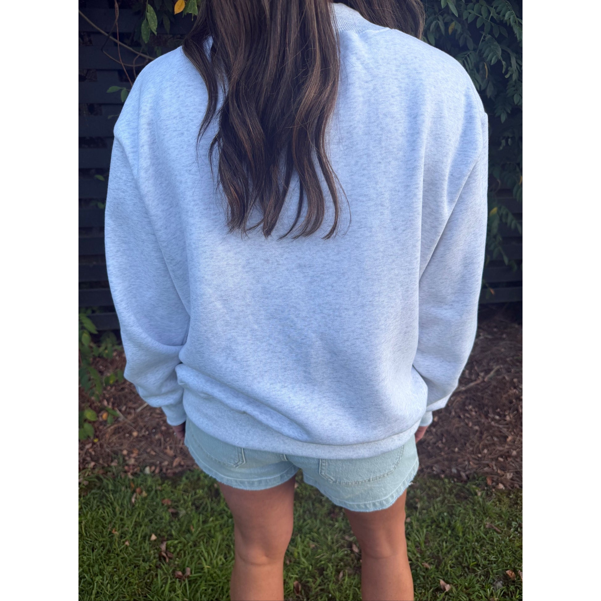 A person viewed from behind wearing a light heather gray crewneck sweatshirt and light blue denim shorts, standing outdoors on grass with a dark fence and plants in the background.
