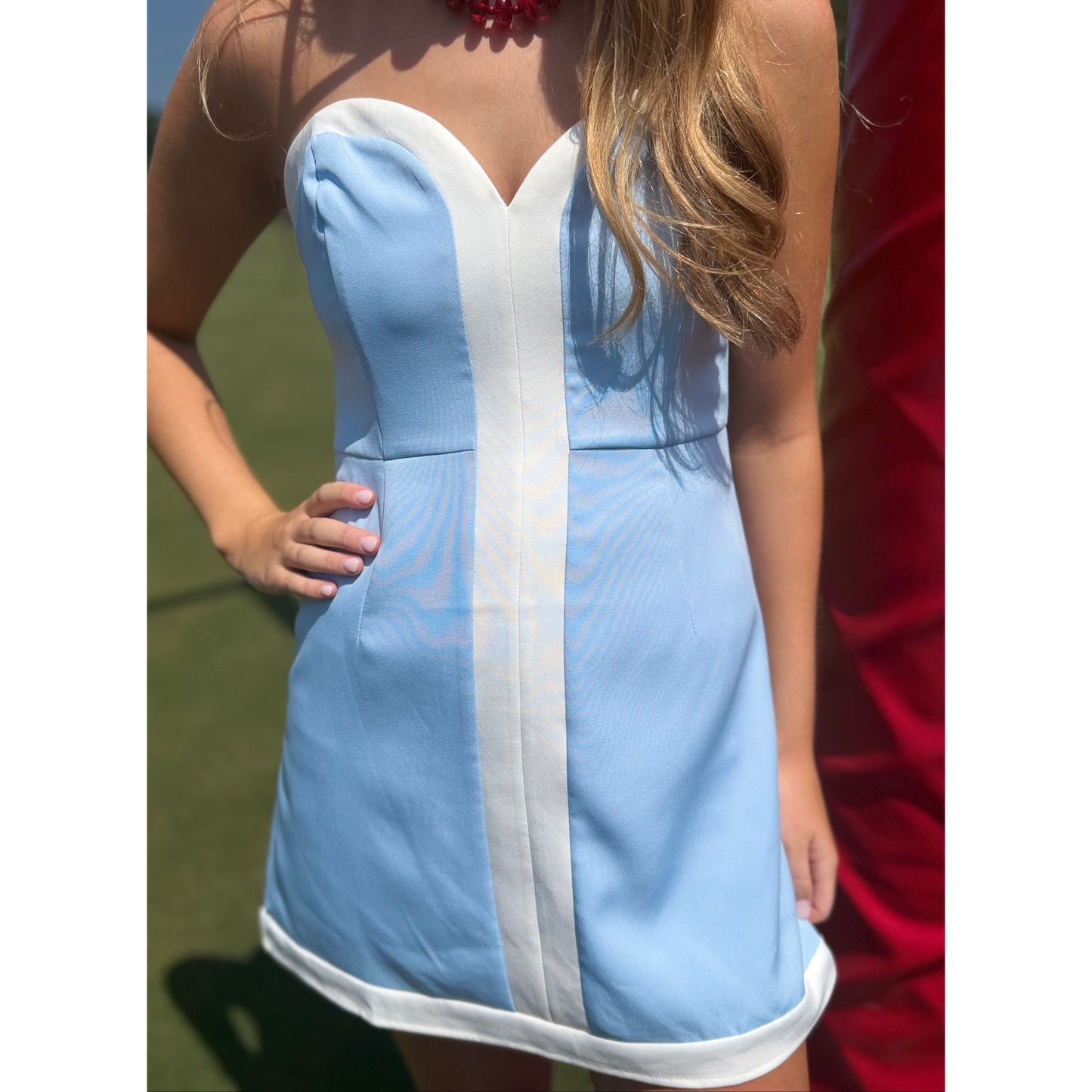 A woman wearing a light blue and white strapless mini dress, photographed outdoors on a sunny day with grass and another person partially visible in the background.