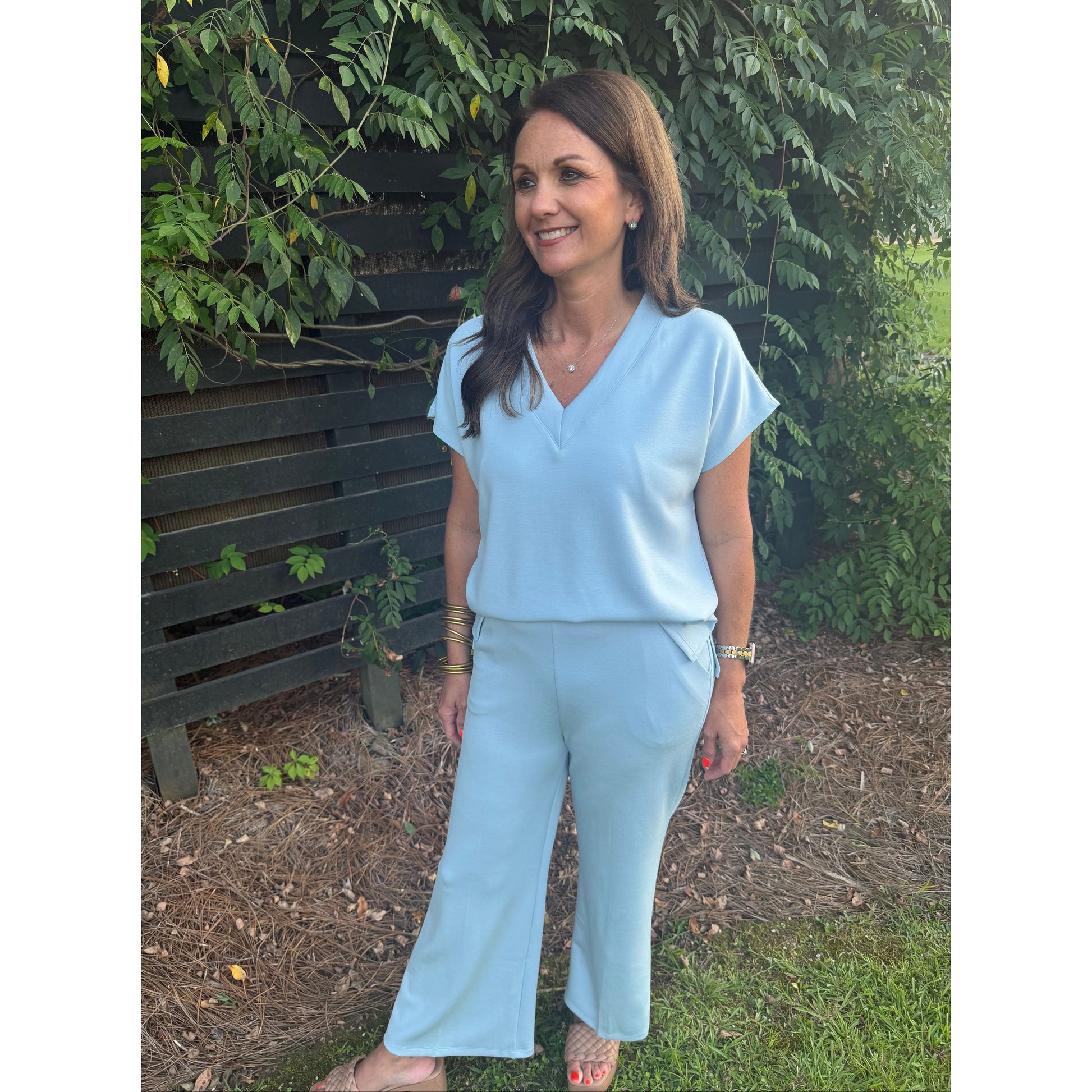 A woman wearing a light blue matching two-piece outfit consisting of a short-sleeve V-neck top and wide-leg pants, standing outdoors on a wooden bench with green foliage and mulch in the background.