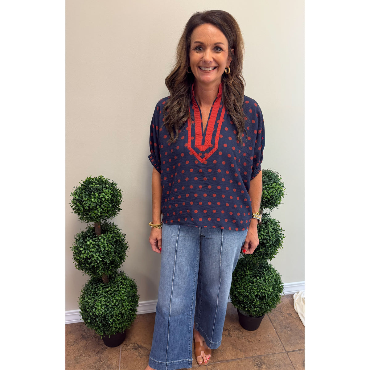A woman stands indoors wearing a navy blue short-sleeve top with red polka dots and a red embroidered V-neckline, paired with wide-leg blue jeans. She is positioned between two green topiary trees against a plain beige wall.