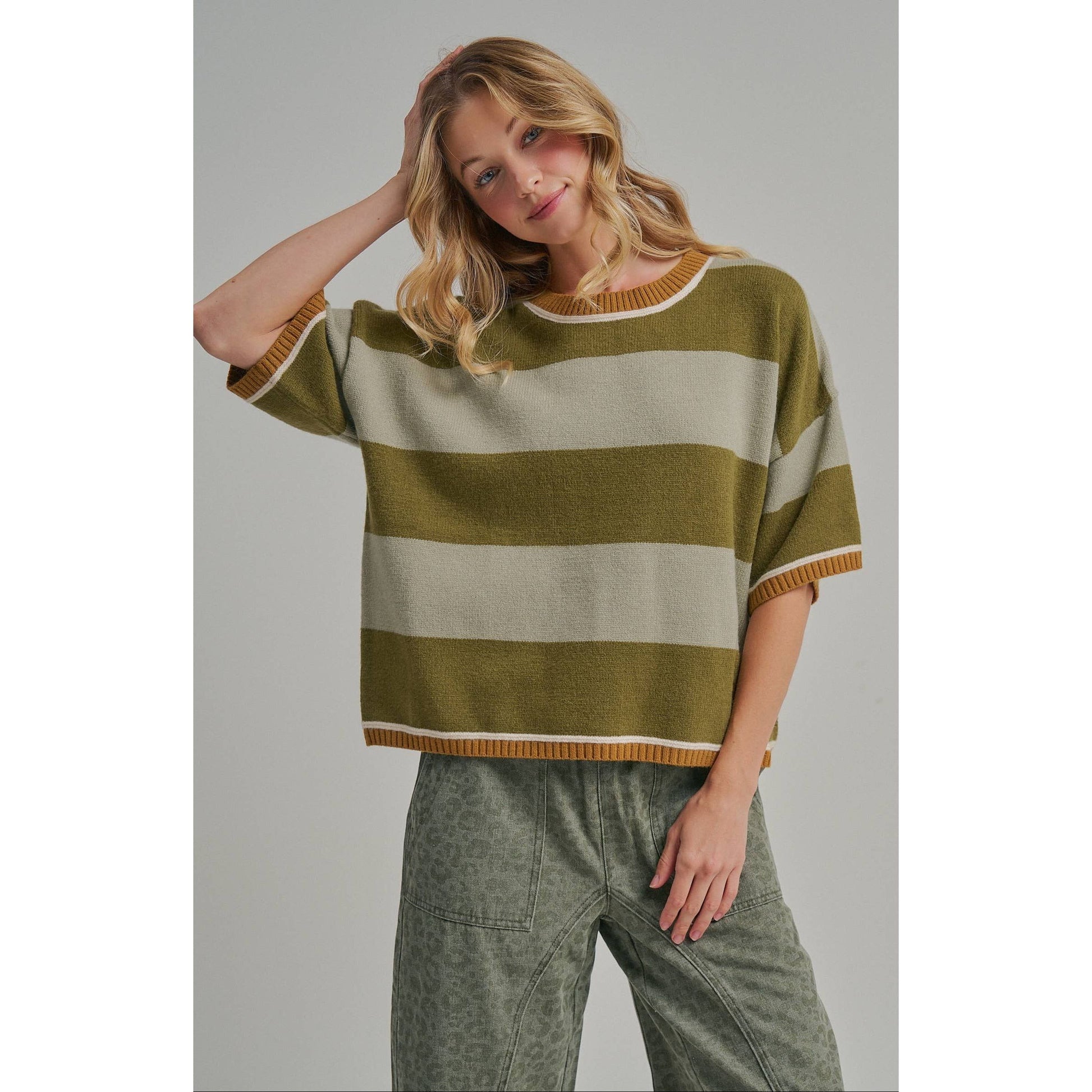 A woman wearing a short-sleeve knit sweater with horizontal olive green and cream stripes, paired with wide-leg gray pants, against a plain light gray studio background.