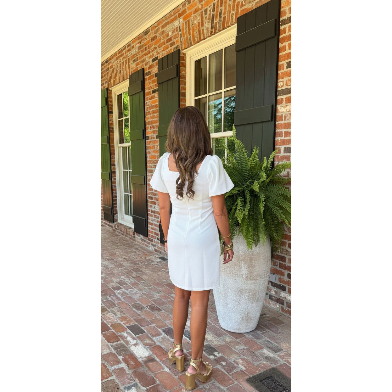Woman in a white dress standing on a brick porch with a plant in the background