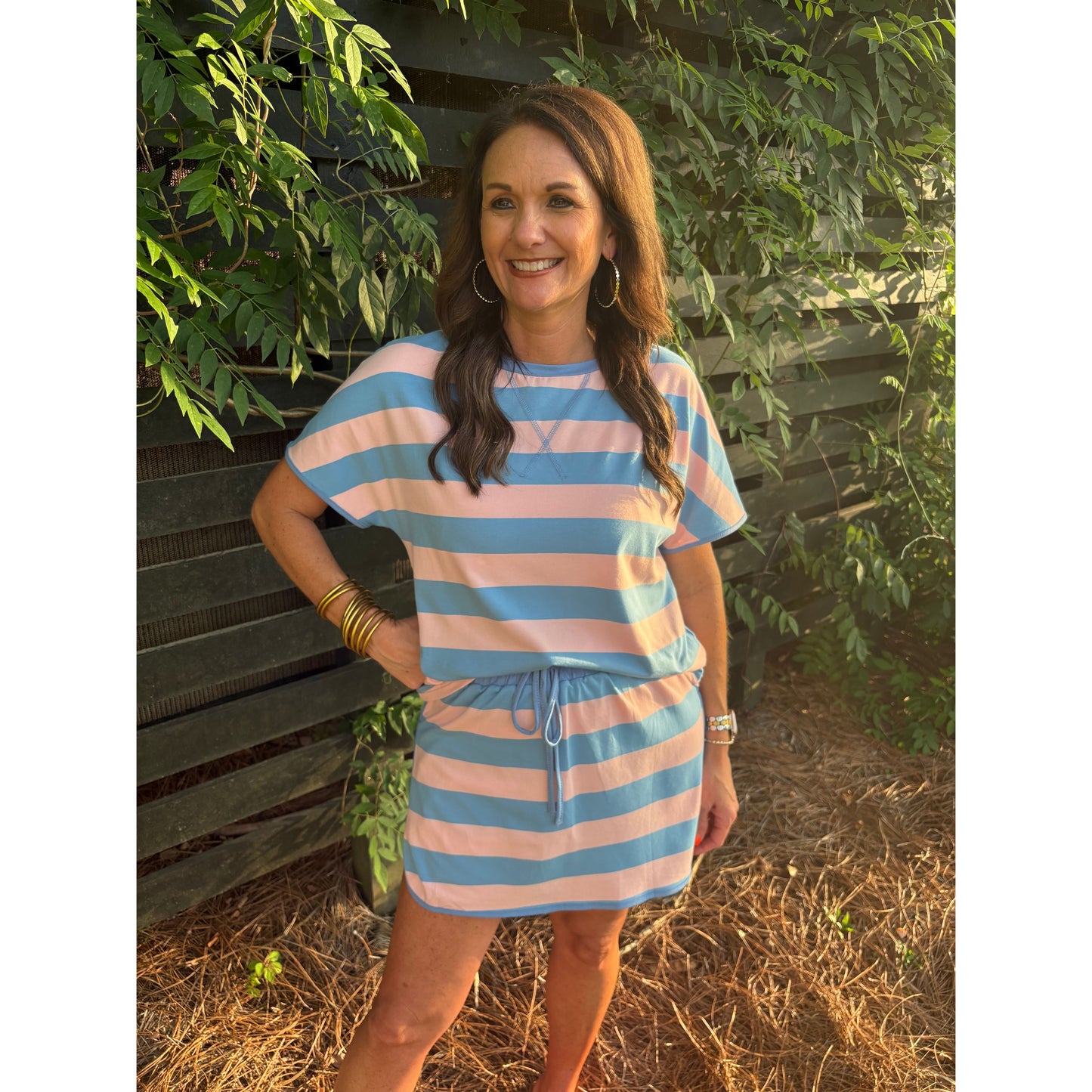 Woman wearing a pink and blue striped dress standing outdoors with greenery in the background